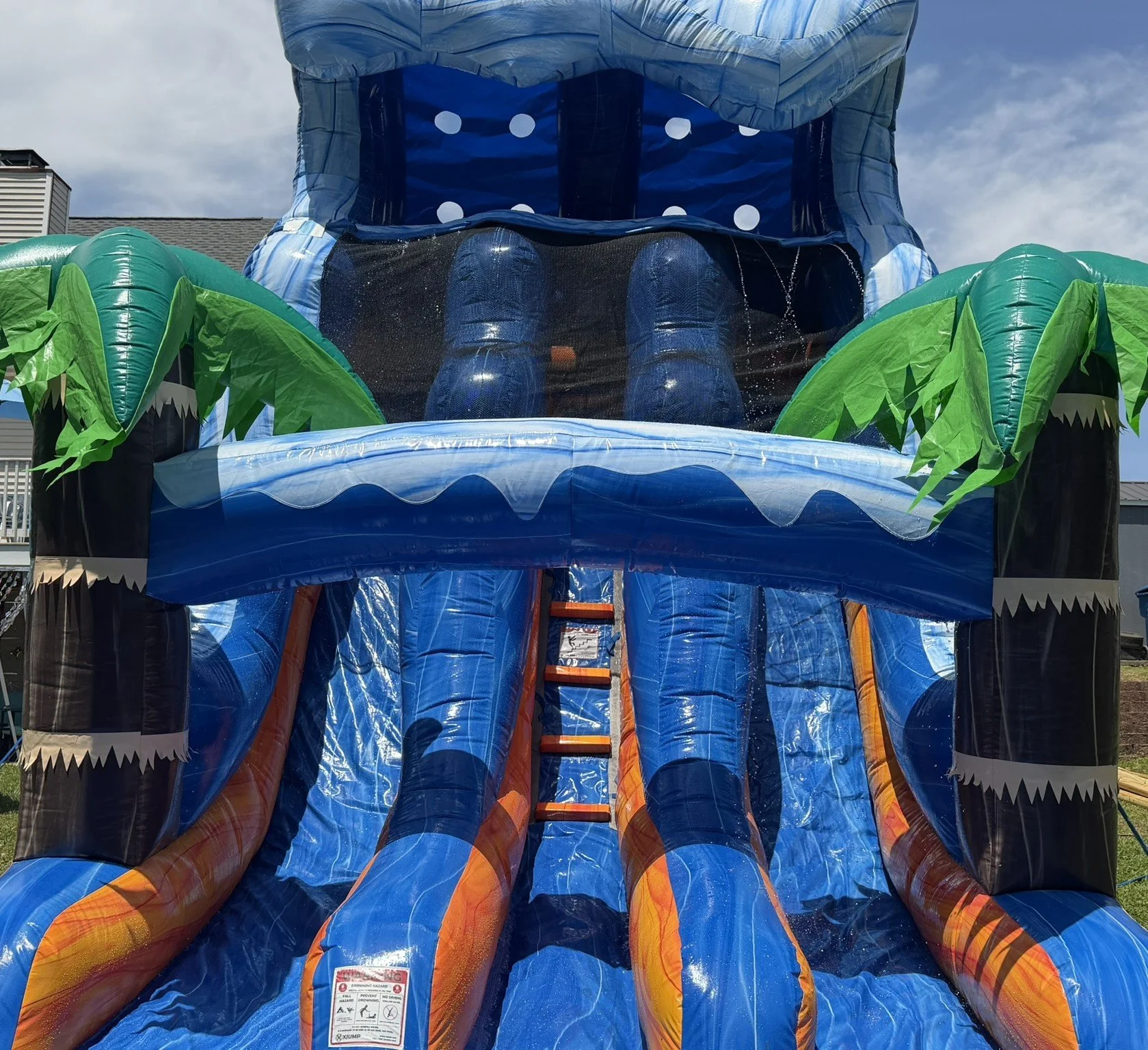 Large inflatable water slide designed to look like a jungle with palm trees and crocodiles, featuring blue, green, orange, and black colors, set outdoors under a cloudy sky.