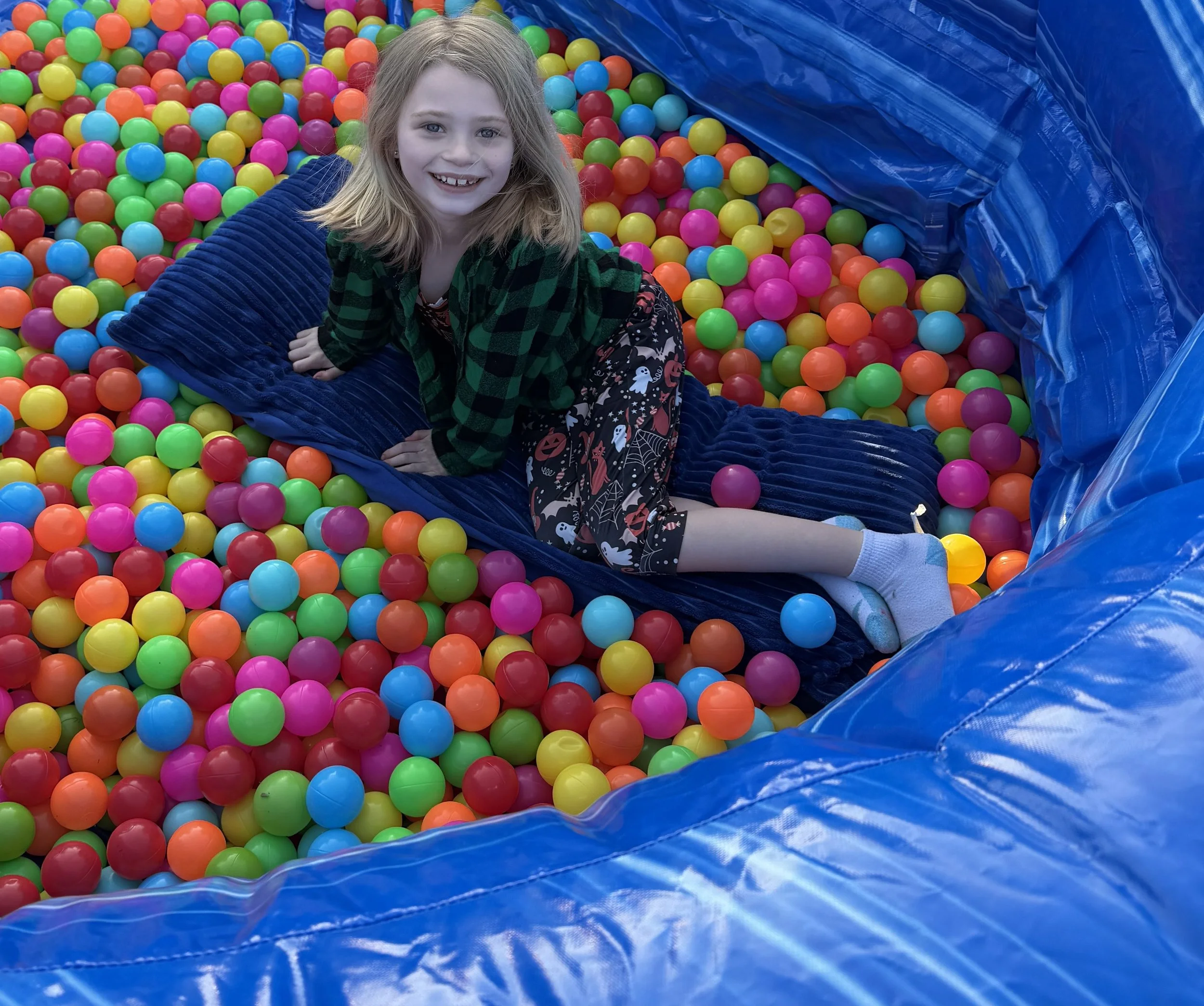 A young girl with blond hair and a big smile, wearing a green and black checkered jacket, sitting in a colorful ball pit filled with red, yellow, green, blue, pink, and orange plastic balls, with a blue inflatable border.