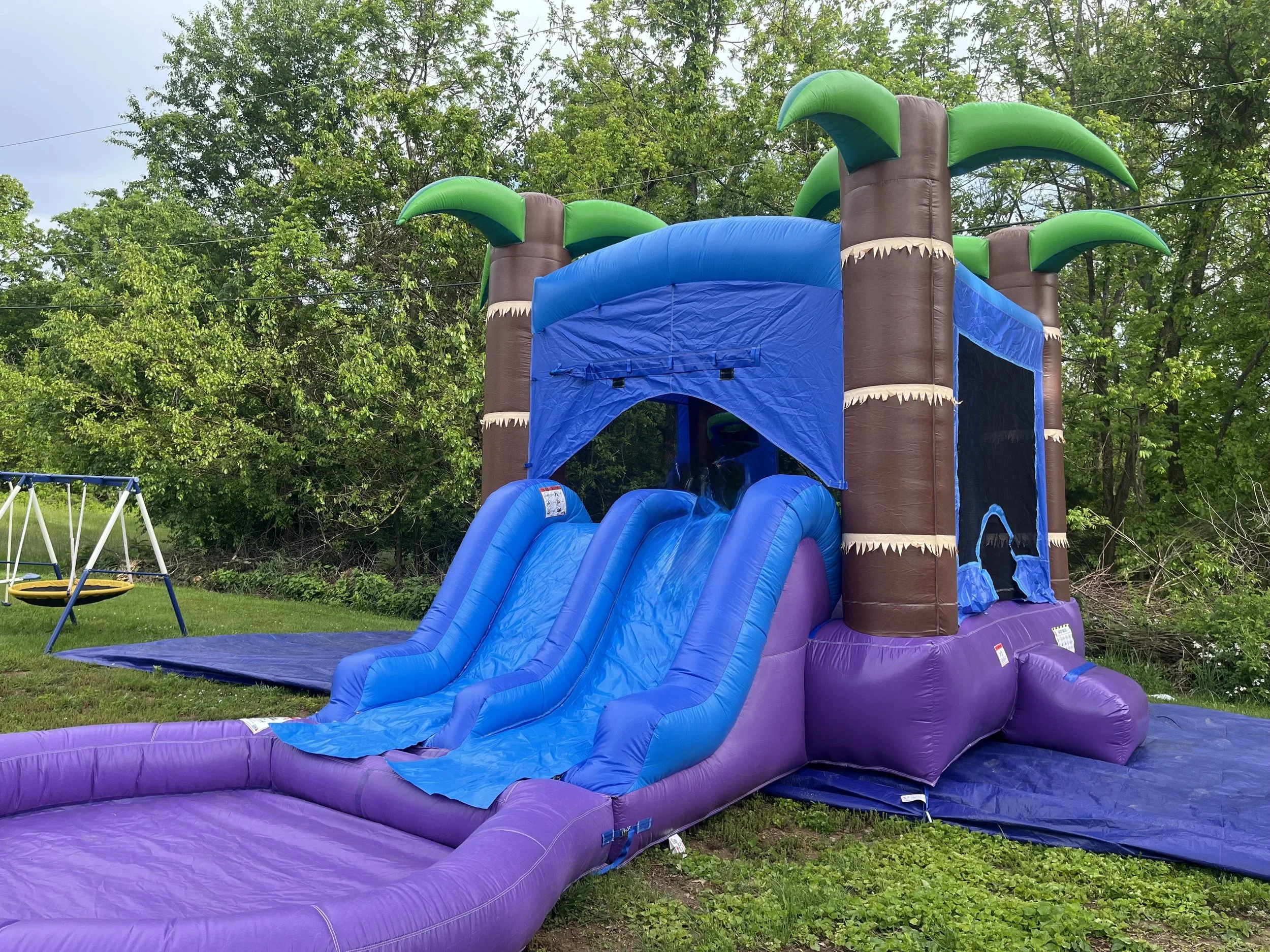 Colorful inflatable outdoor water slide with a blue slide portion and palm tree decorations at the top, set in a backyard with green trees and a swing set nearby.