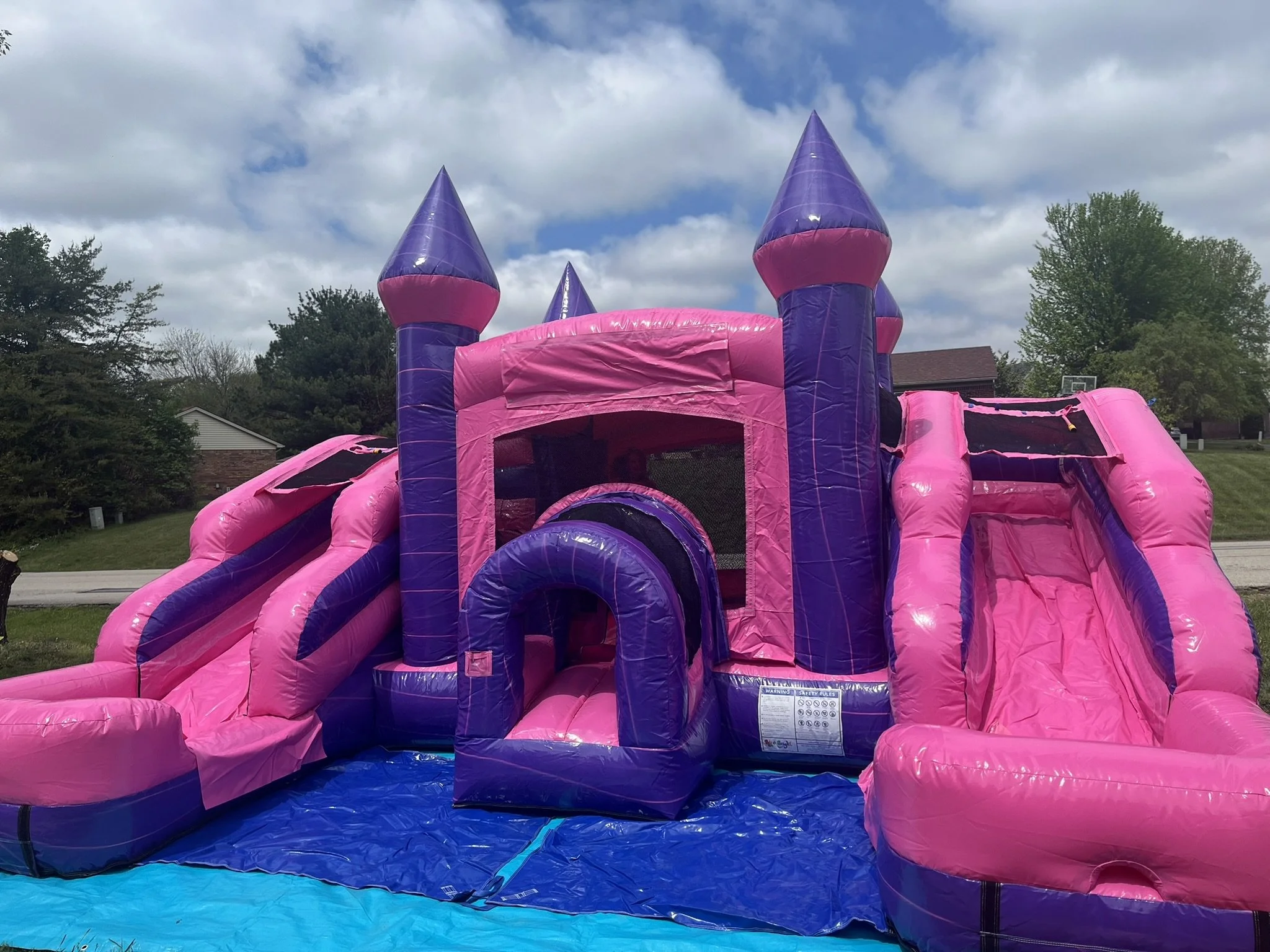 Pink and purple inflatable bounce house resembling a castle with towers and slide, set outdoors under cloudy sky.