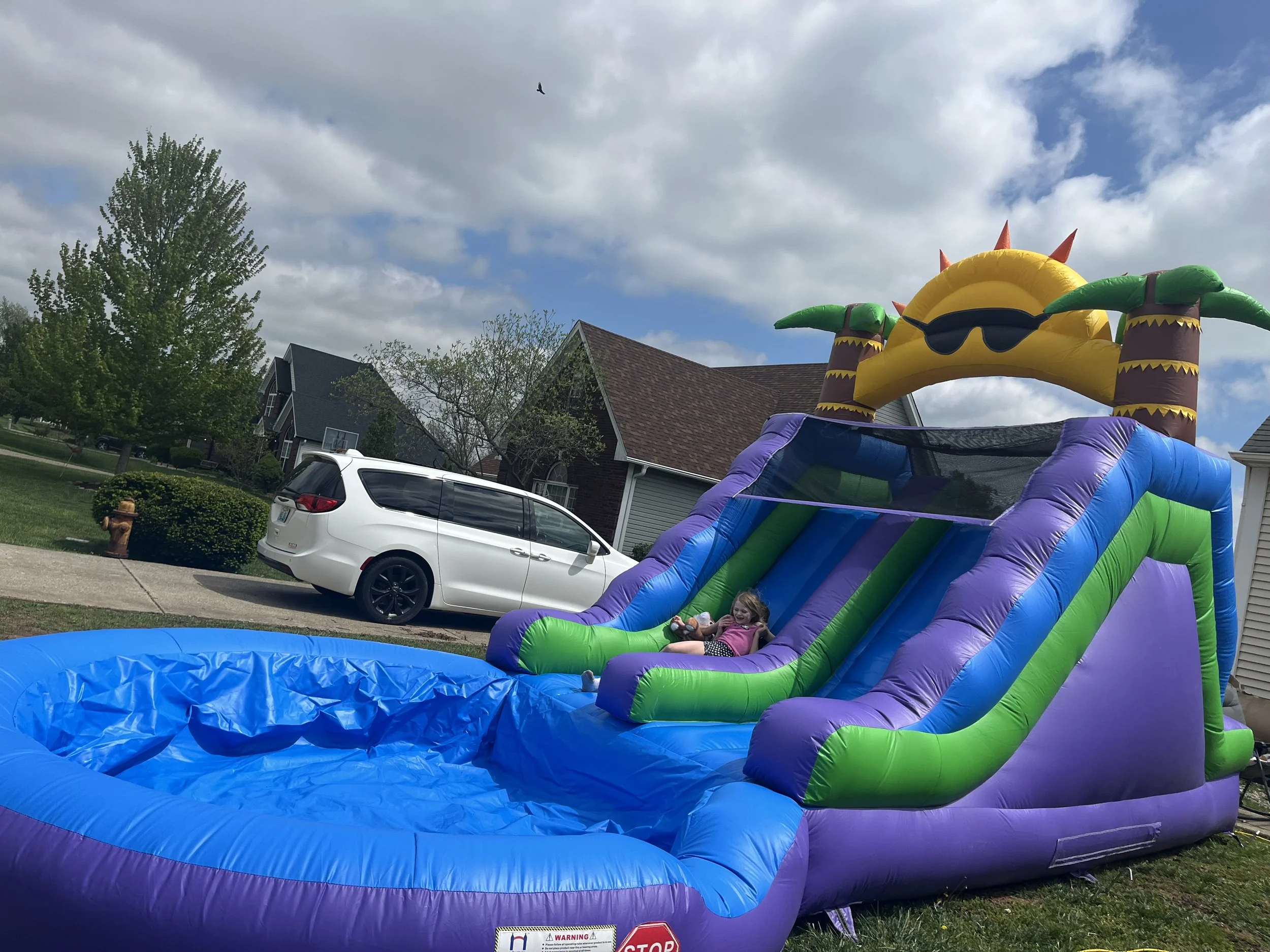 Two children, a girl and a boy, are sliding down on a colorful inflatable slip and slide in a suburban backyard, with houses and trees in the background.