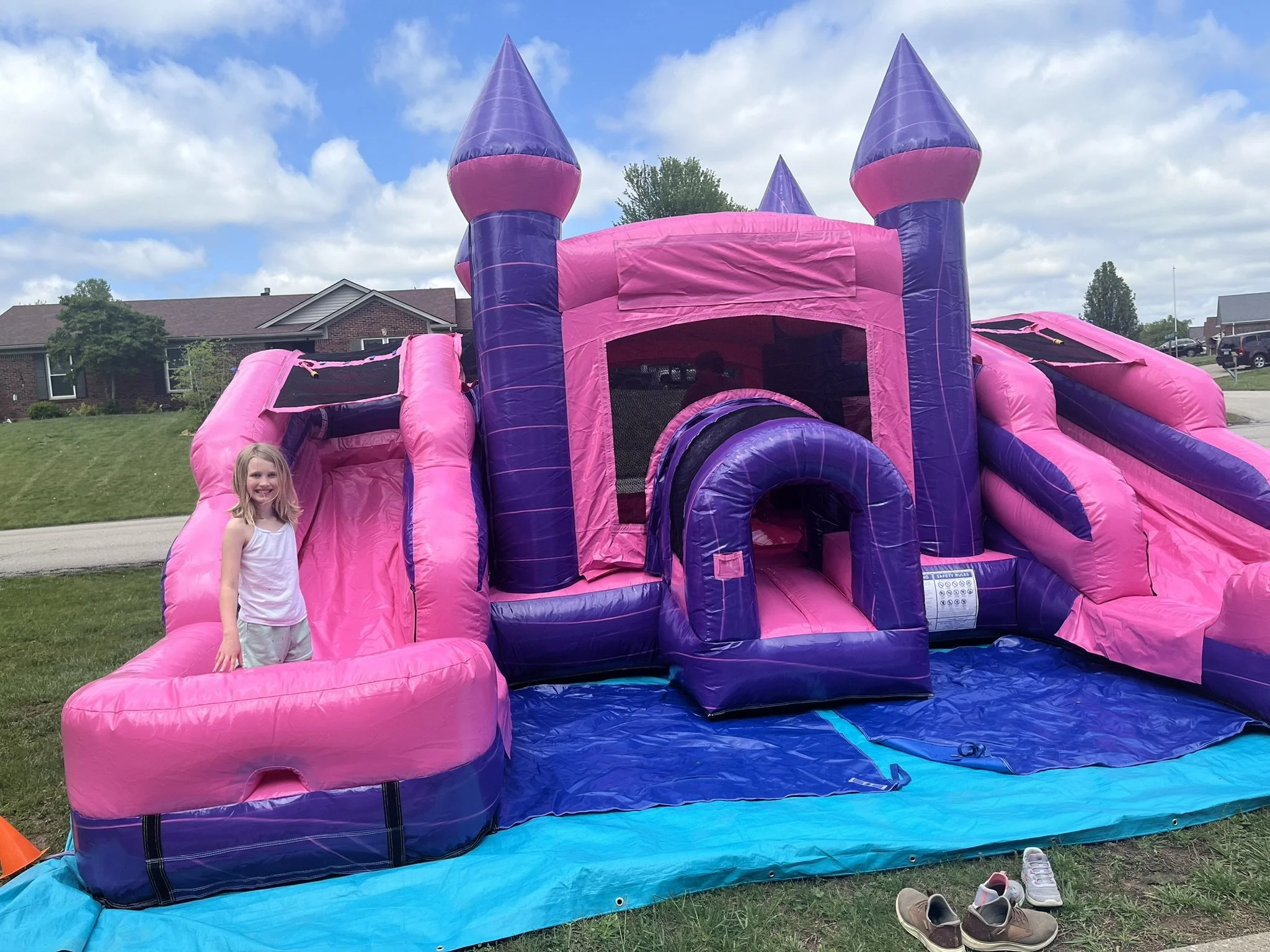 A pink and purple inflatable bounce house designed like a castle with a slide, set up outdoors on a grassy area. There is a young girl with blonde hair and a white shirt standing next to it, smiling. The sky is partly cloudy, and houses are visible in the background.