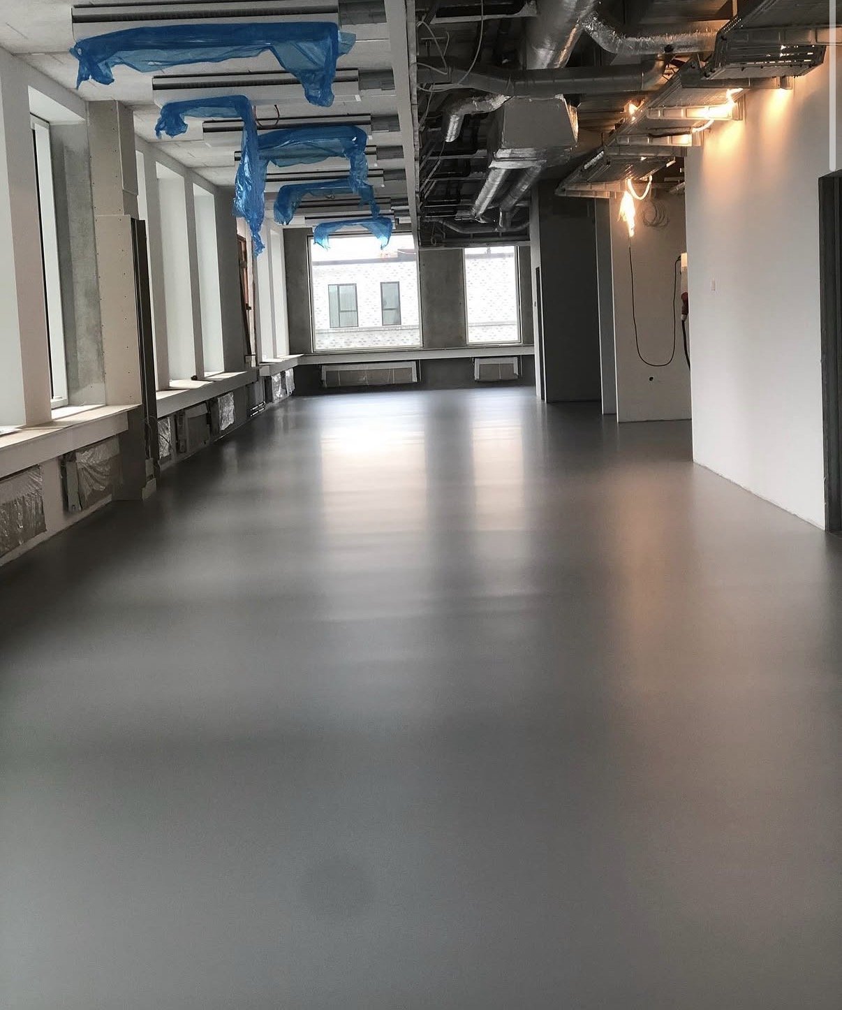 Et tomt kontorlokale under renovering med store vinduer, udsugningssystemer i loftet og midlertidig plastikdække på dele af loftet.