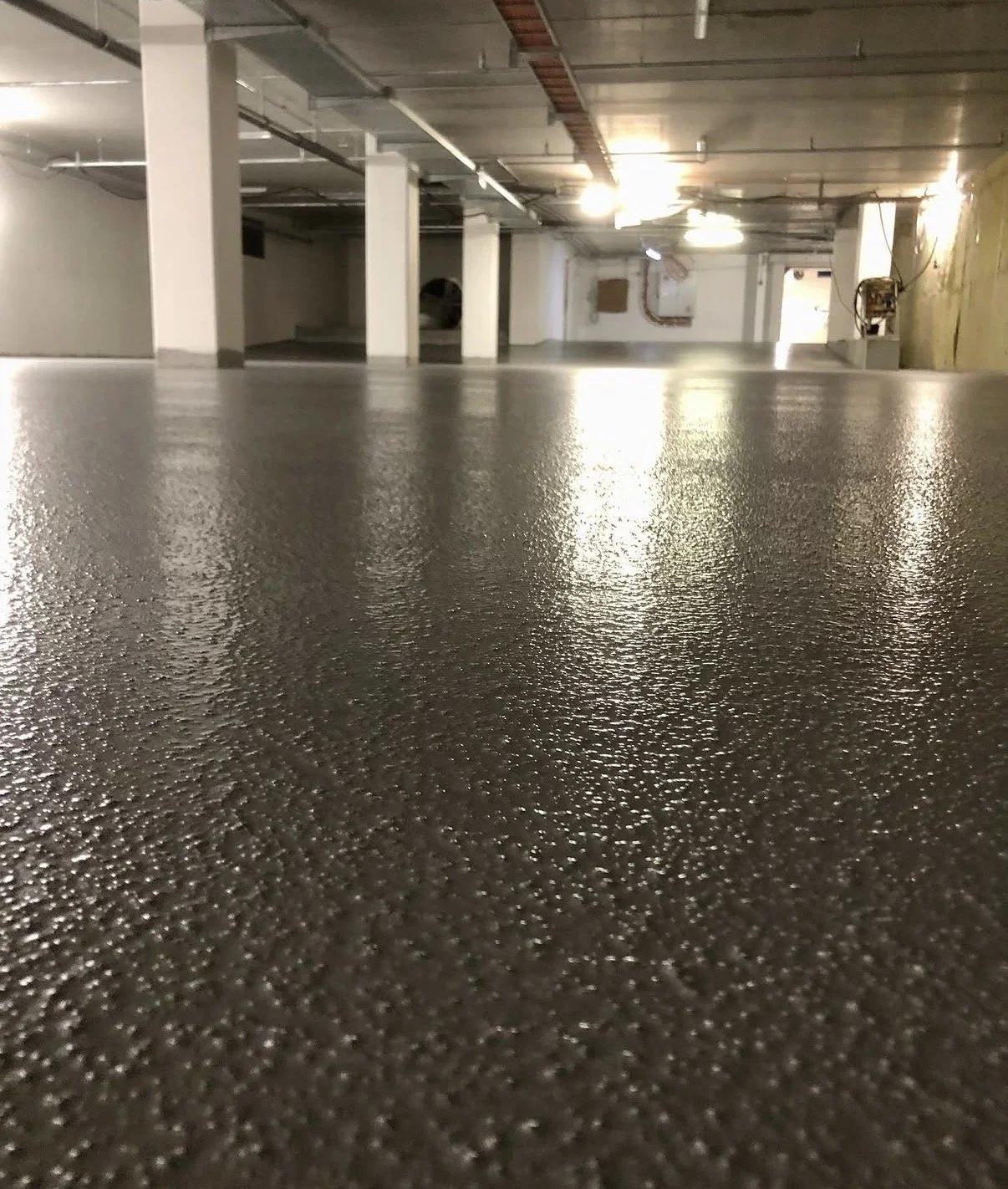 Klipset, betonlignende gulv i en underjordisk parkeringskælder med hvide støttepiller og synlige rør i loftet.
