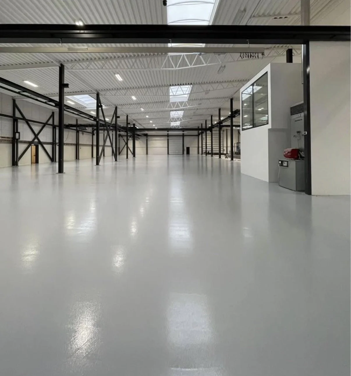 Et stort, tomt lager med hvid polished gulv, hvide vægge og metalbjælker, med flere skylights i loftet. Der er en kontorbygning i den ene side med et vindue.