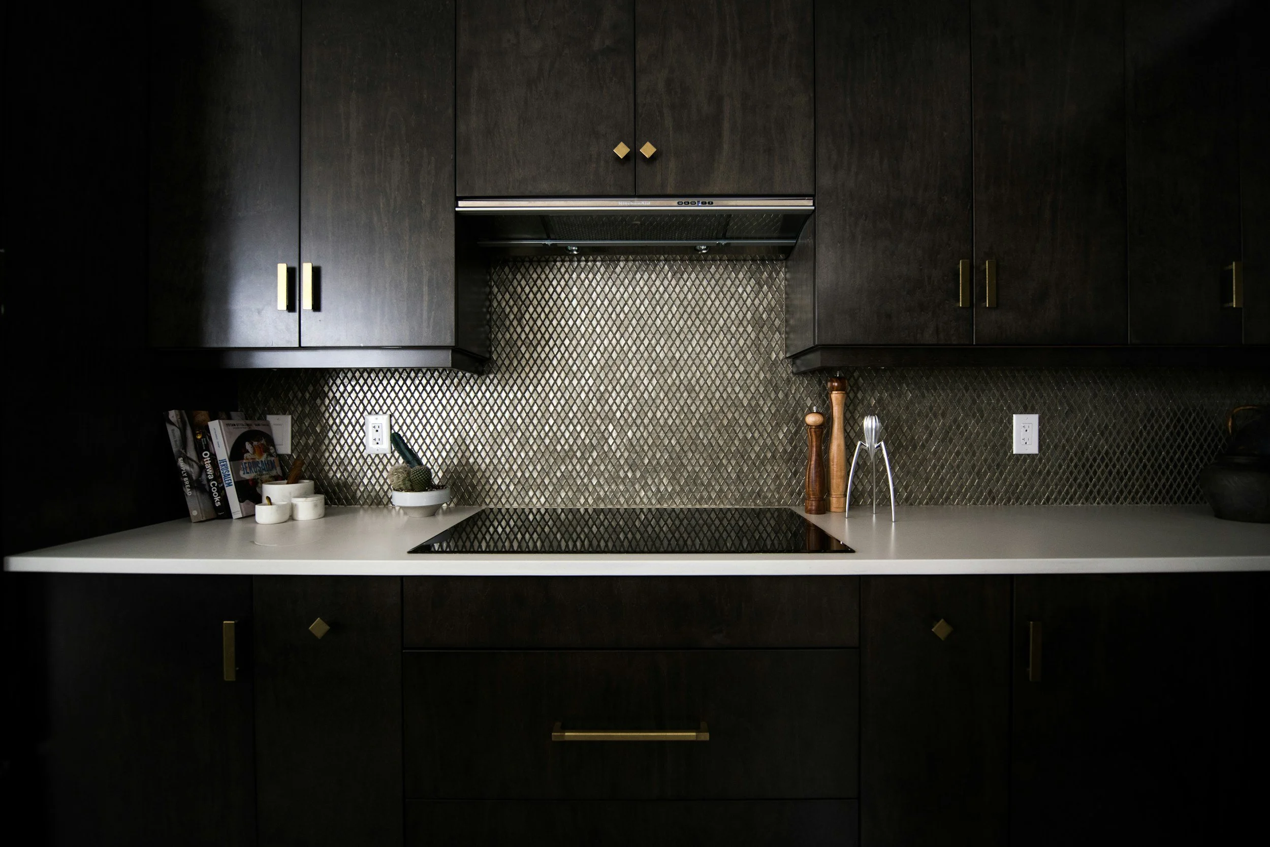 Dark kitchen cabinets above a white countertop with a black stovetop. Items on the countertop include cookbooks, a bowl, salt and pepper shakers, and kitchen utensils against a patterned backsplash.