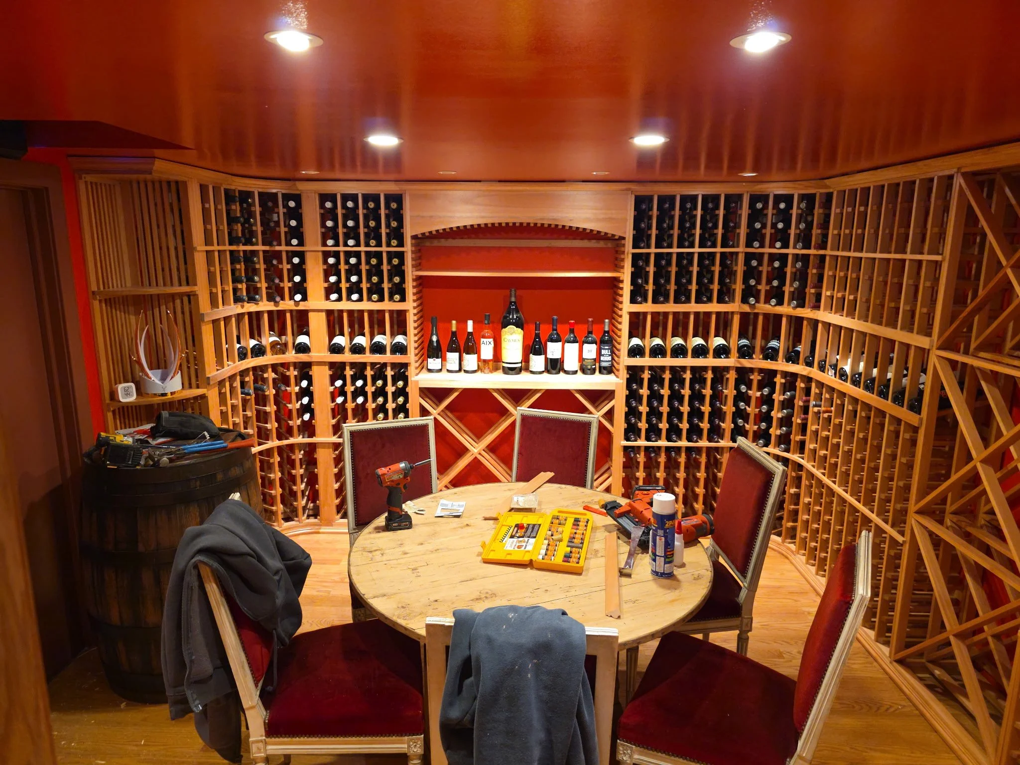 A wine cellar with wooden shelves filled with wine bottles, a table with tools and supplies, chairs around the table, and ceiling lights overhead.