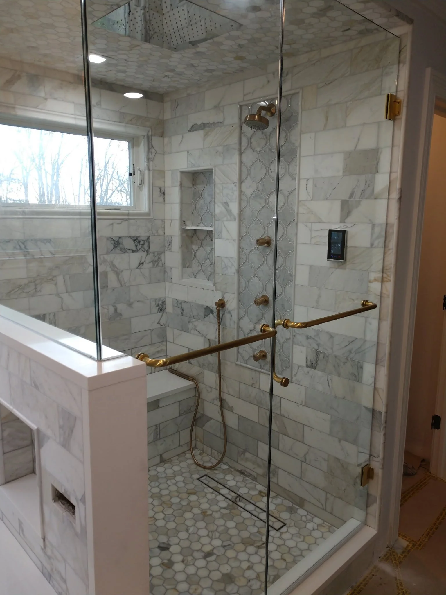 A glass shower enclosure with brass fixtures, marble tile walls with decorative accents, a window, and a built-in shelf.