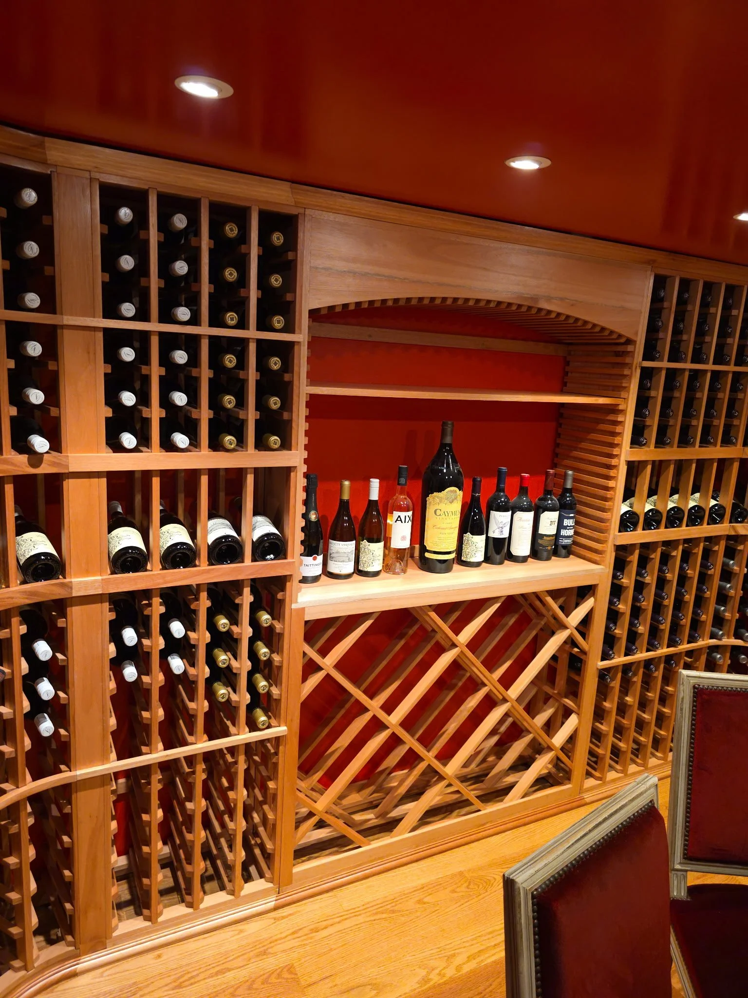A wine cellar with wooden shelves filled with bottles of wine and a countertop displaying more bottles. Red chairs are visible in the foreground.