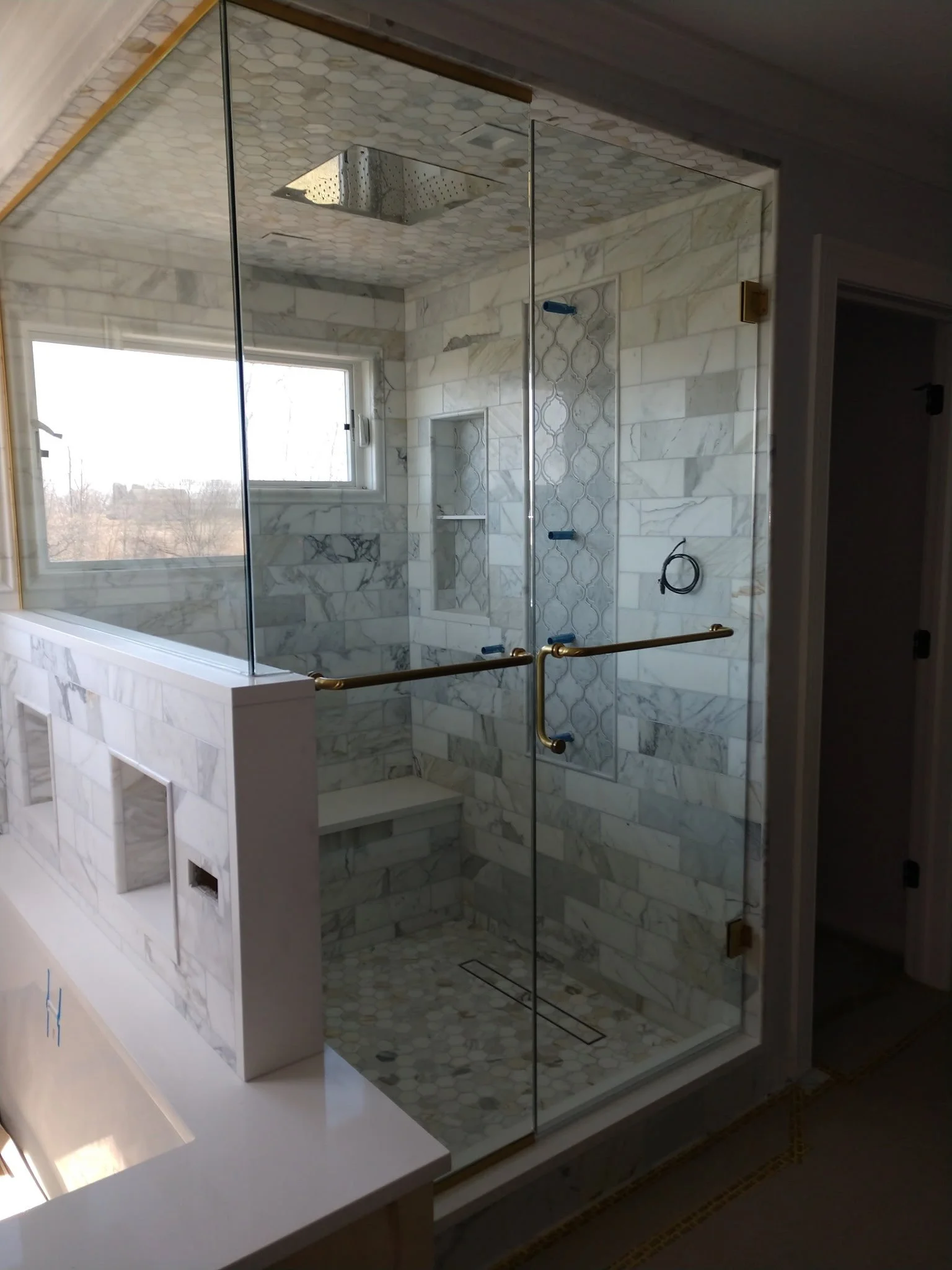 A modern walk-in shower with glass doors, tiled with white and gray marble, a window, built-in shelves, and a rain shower head on the ceiling.
