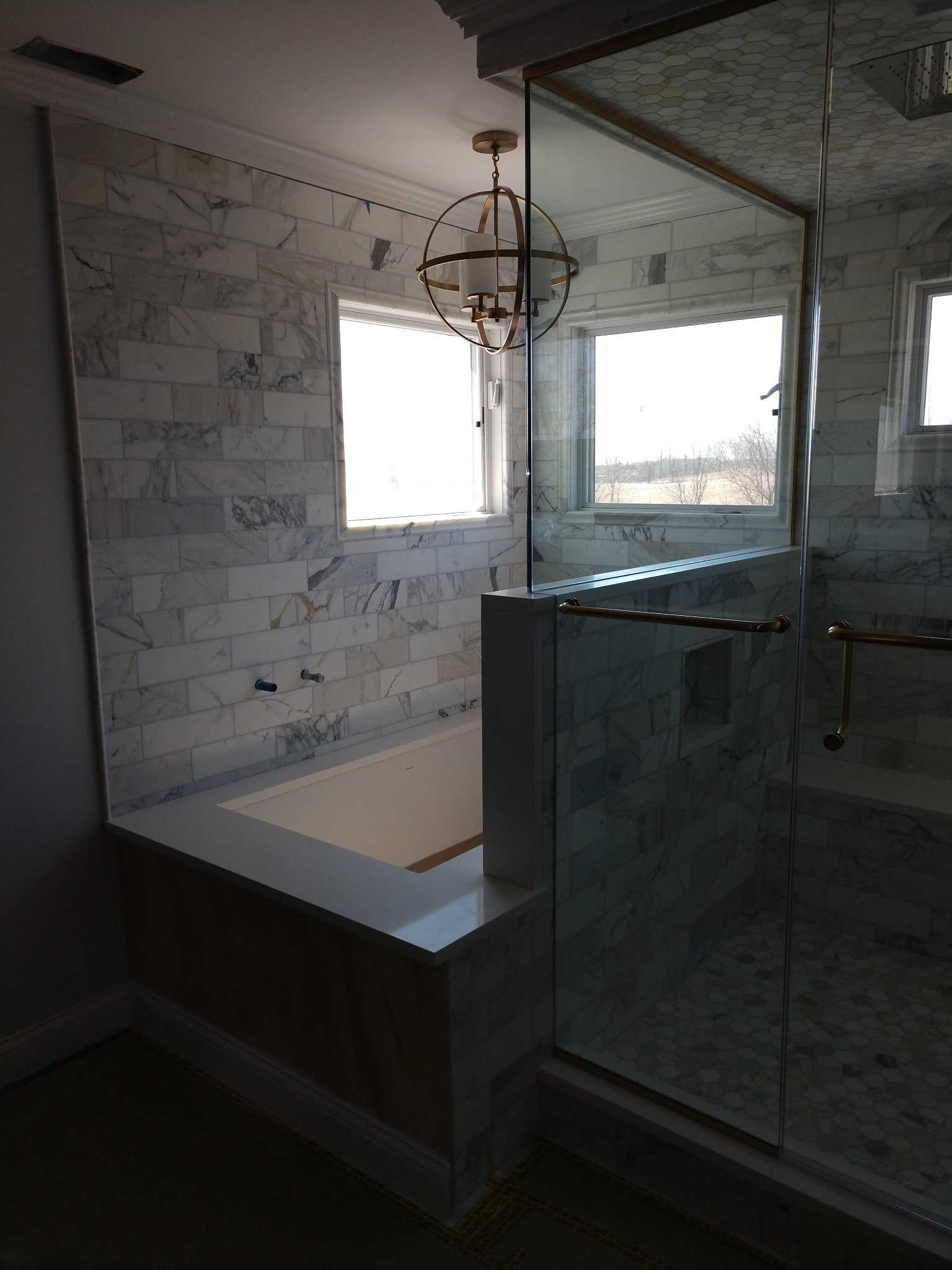 Bathroom with marble walls, a window, a chandelier light fixture, an empty bathtub, and a glass-enclosed shower with marble tiles and brass handles.