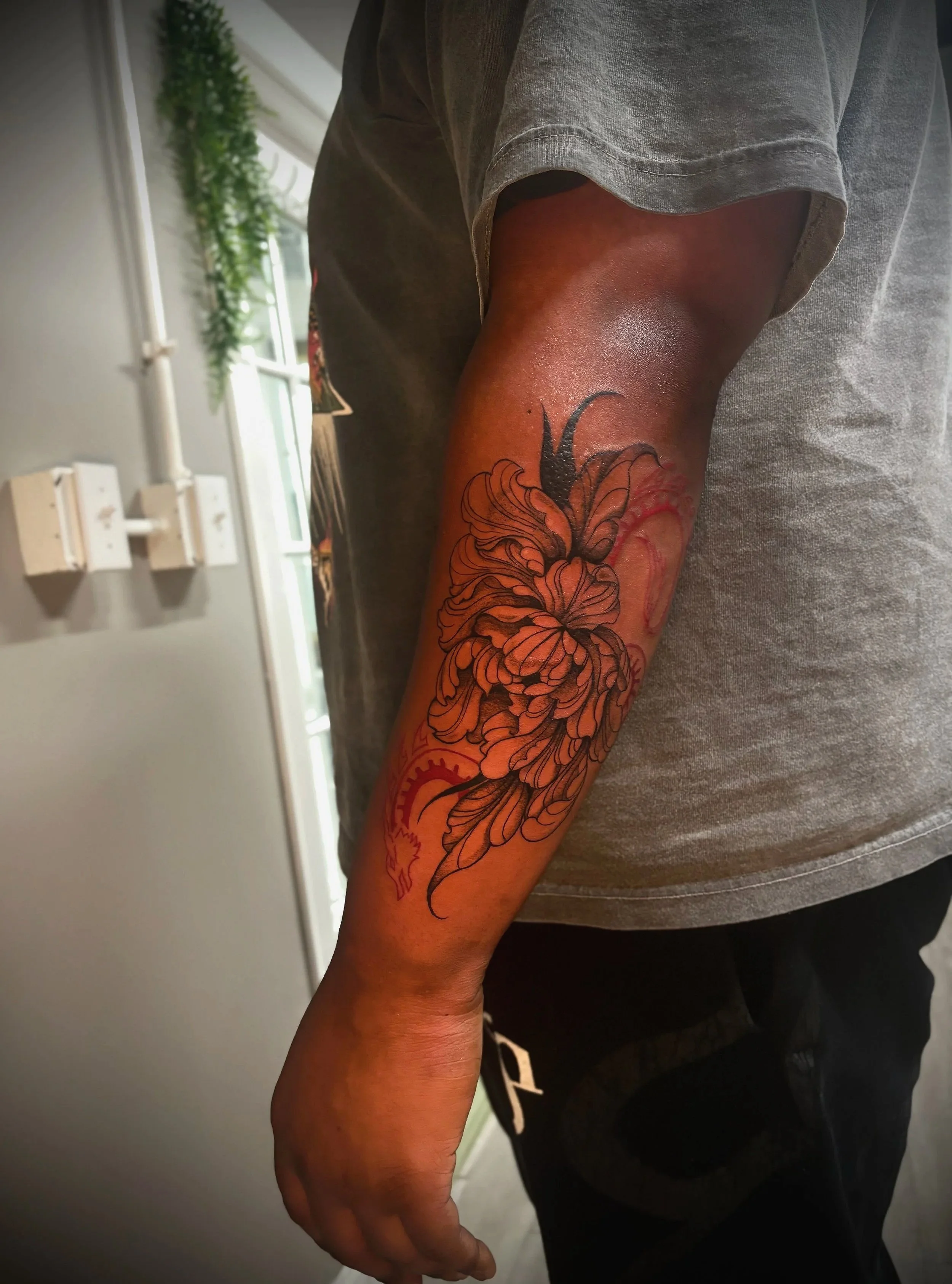 A person's arm with a colorful floral tattoo on the forearm, wearing a gray t-shirt, in a room with a window, some greenery, and electrical outlets on the wall.