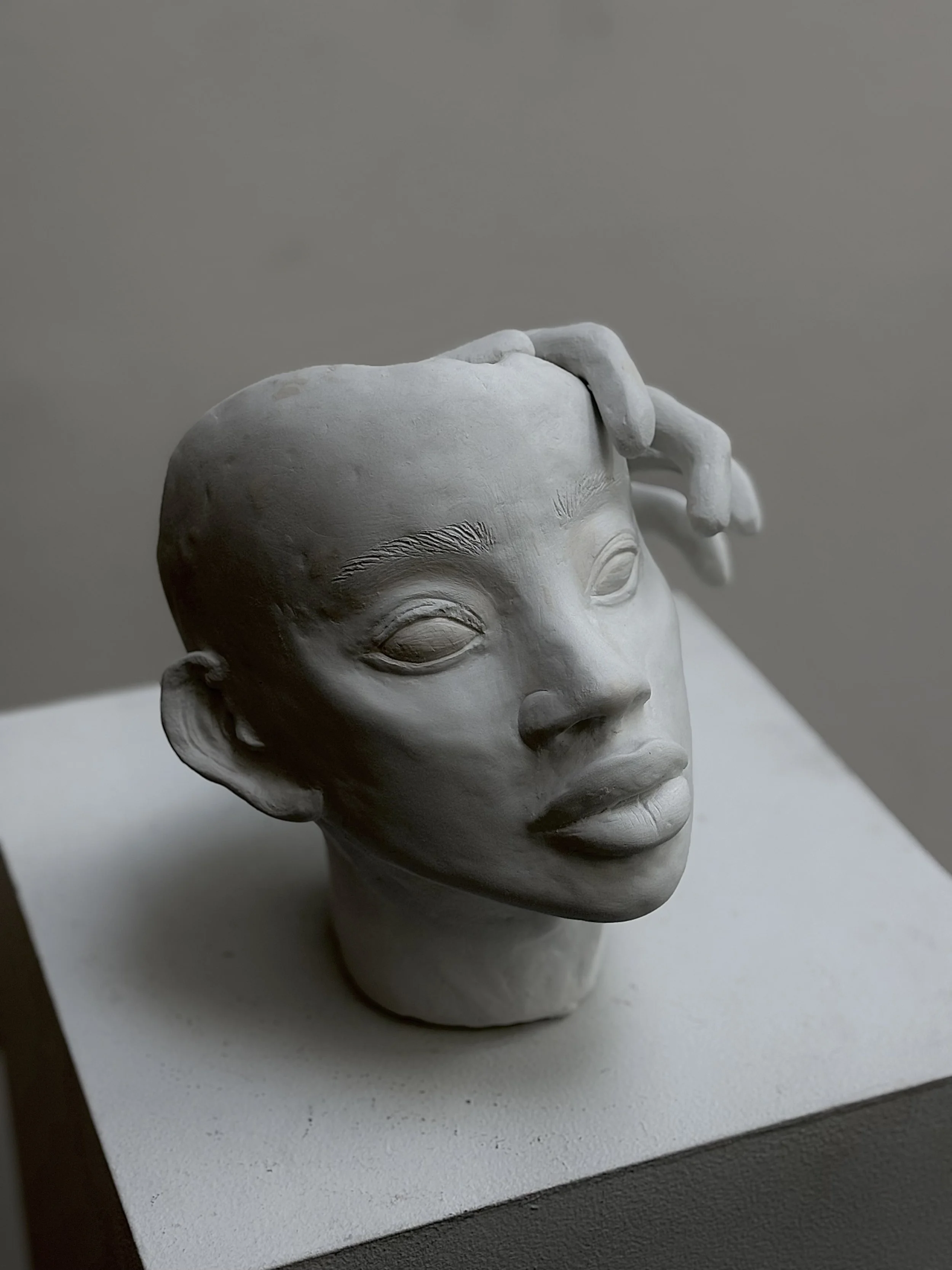 A white sculpture of a woman with closed eyes, short hair, and a hand resting on her head, displayed on a white pedestal against a plain gray background.