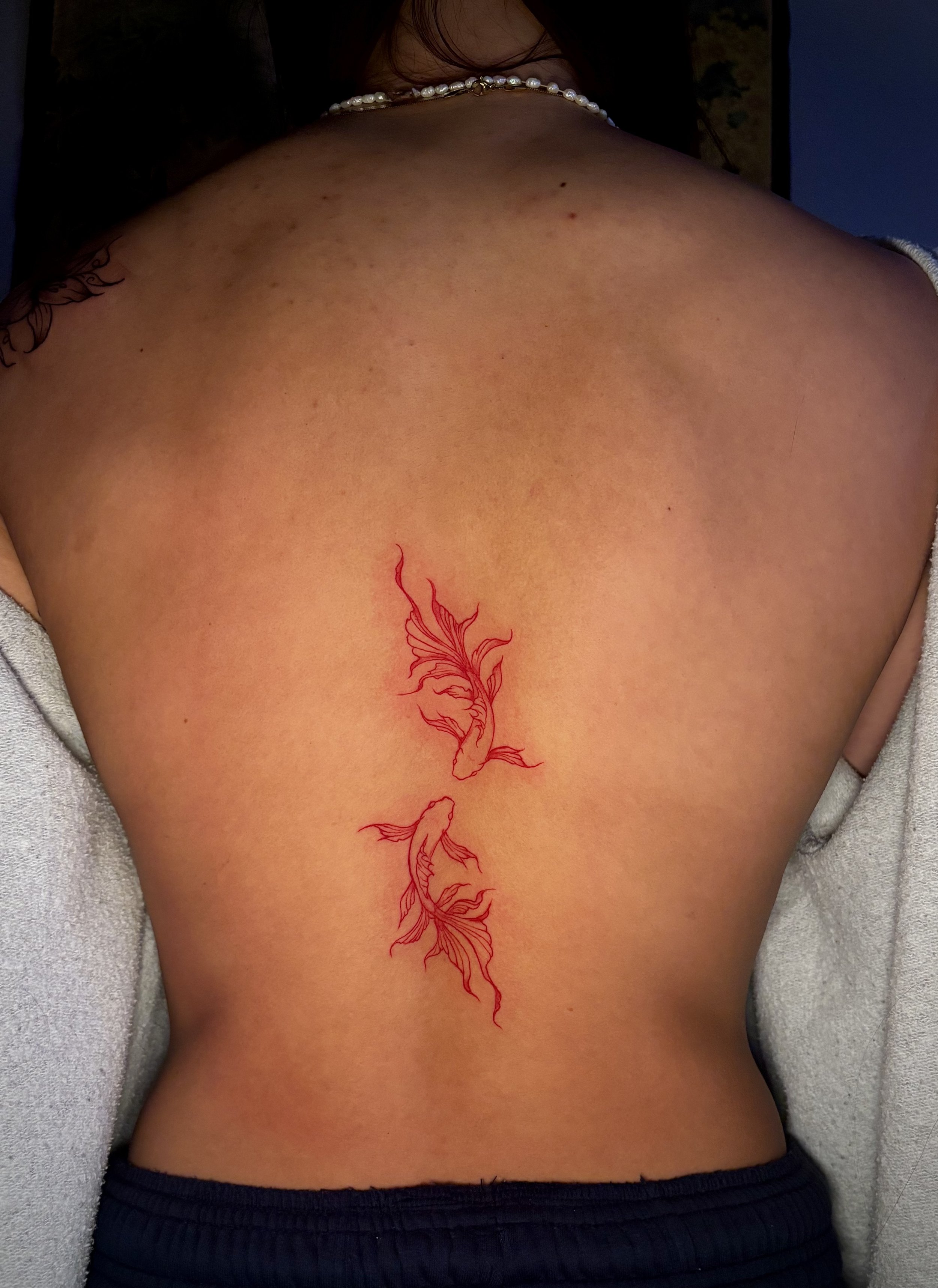 Back of a person with a red tattoo of a koi fish on the lower back, wearing a pearl necklace and a white towel around the shoulders.