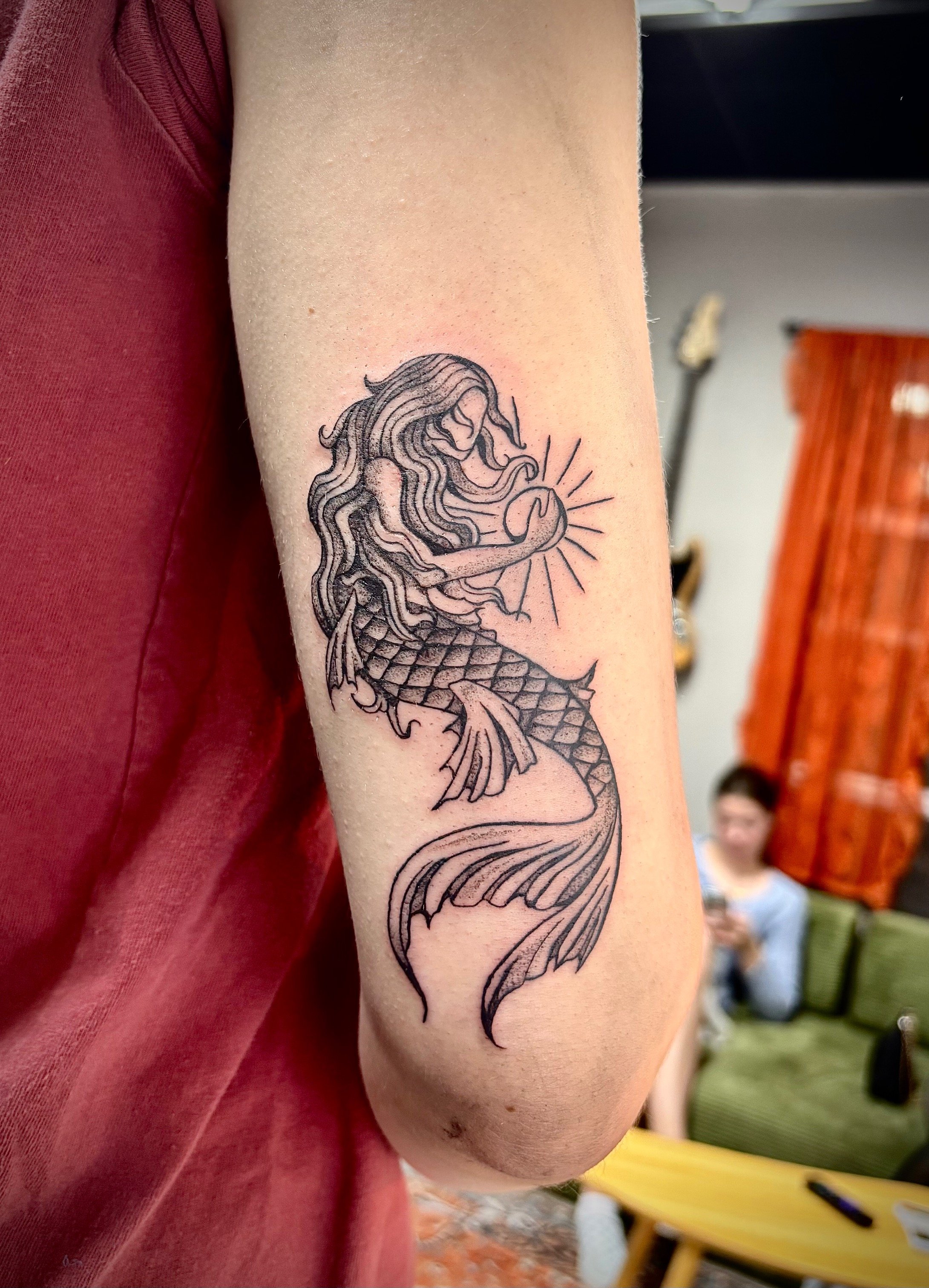 A person's upper arm with a detailed black and gray tattoo of a mermaid holding a sun, with the mermaid's long hair flowing and her tail curling downward.