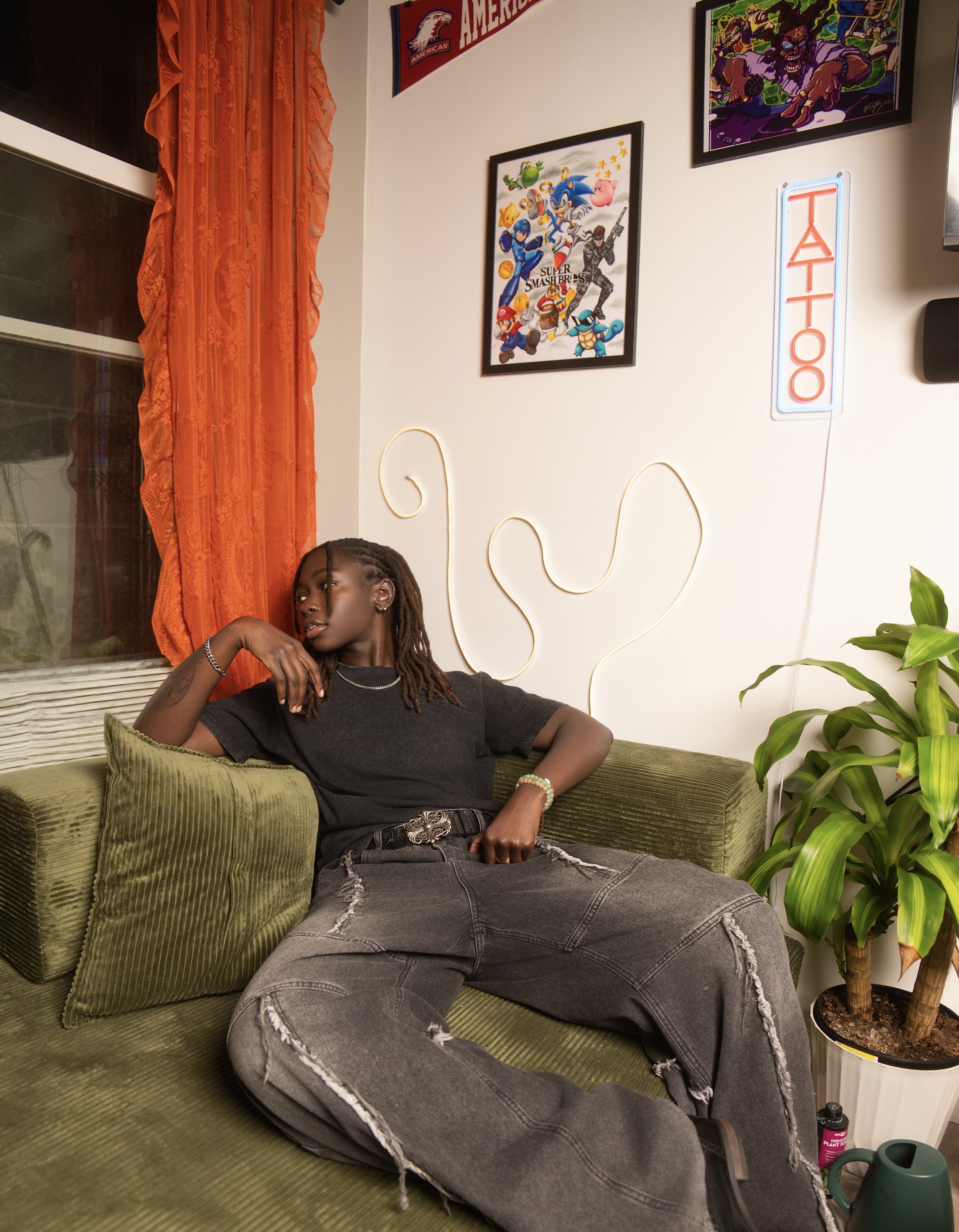 A young woman with dreadlocks reclining on a green velvet sofa in a cozy room decorated with posters and neon signs, including a neon sign that says 'TATTOO'. Black owned business. Queer owned business. Small business. Clean tattoo studio.