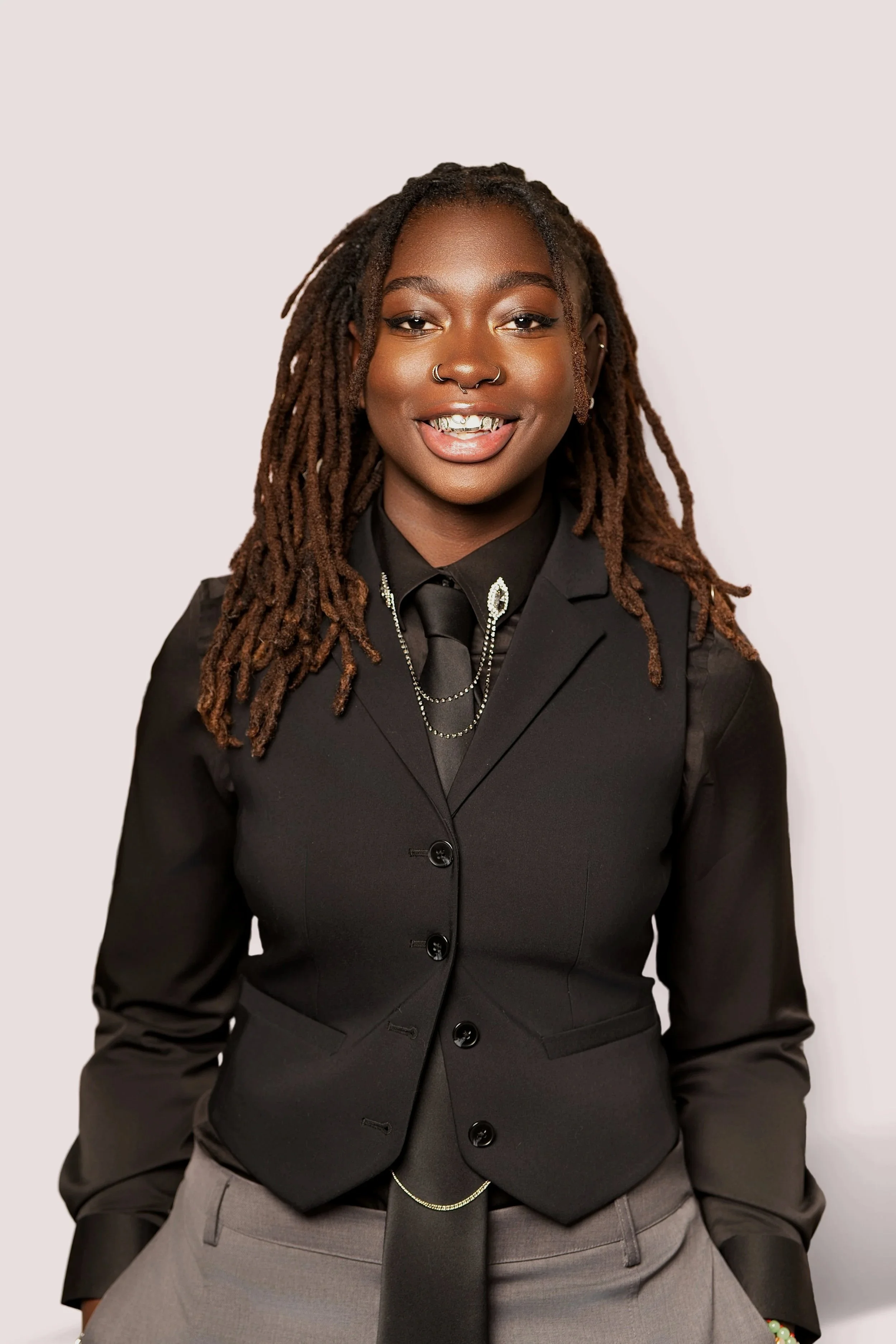 A woman with dark skin in a black blazer, gray pants, and a black tie, smiling, with dreadlocks, jewelry, and a pierced nose, standing against a plain light background. Black, queer, and female business owner.