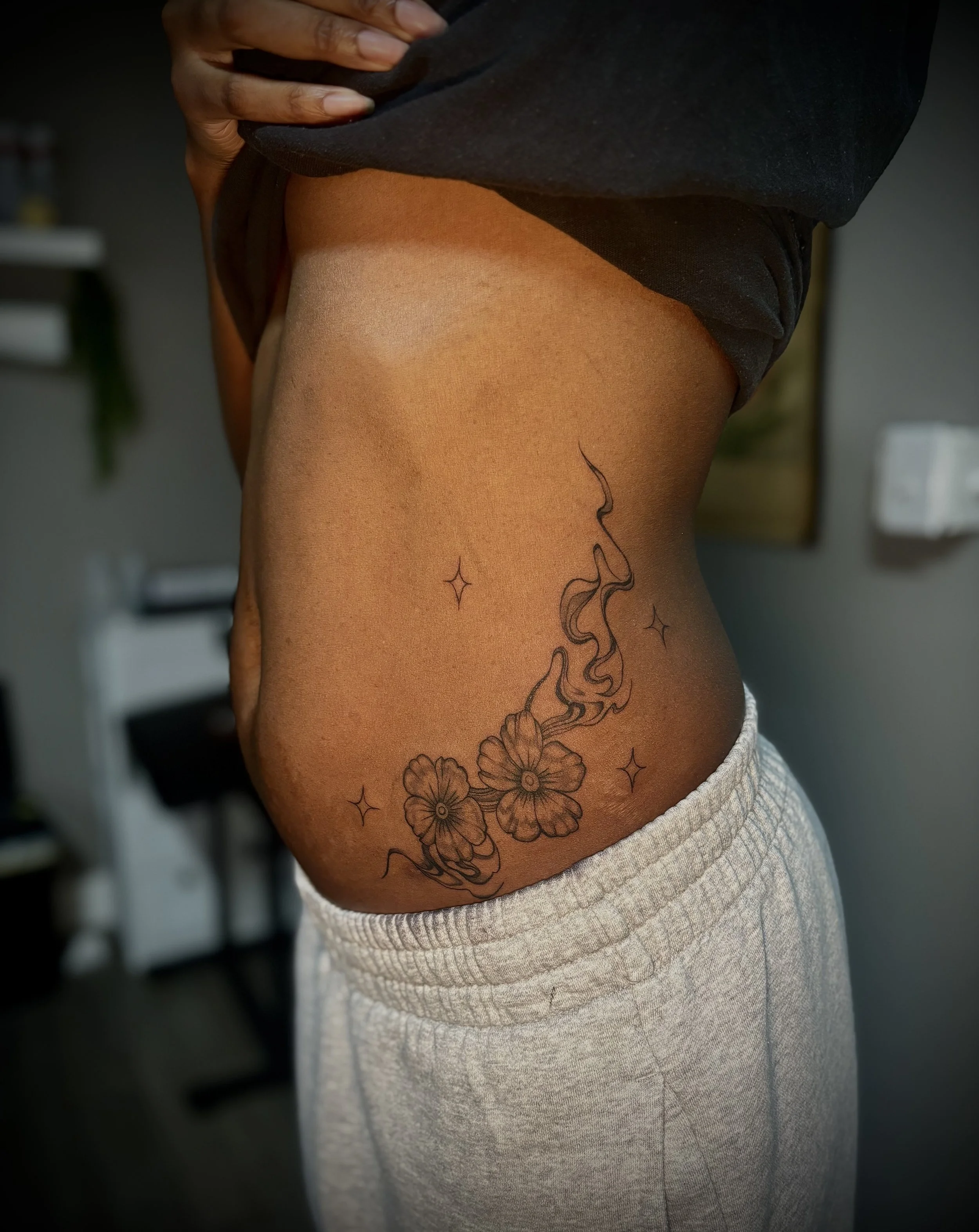Close-up of a person's side, showing a floral tattoo with flowing lines on the ribcage and a hand gently pulling up a dark shirt to reveal it. The person is wearing light-colored, textured sweatpants.