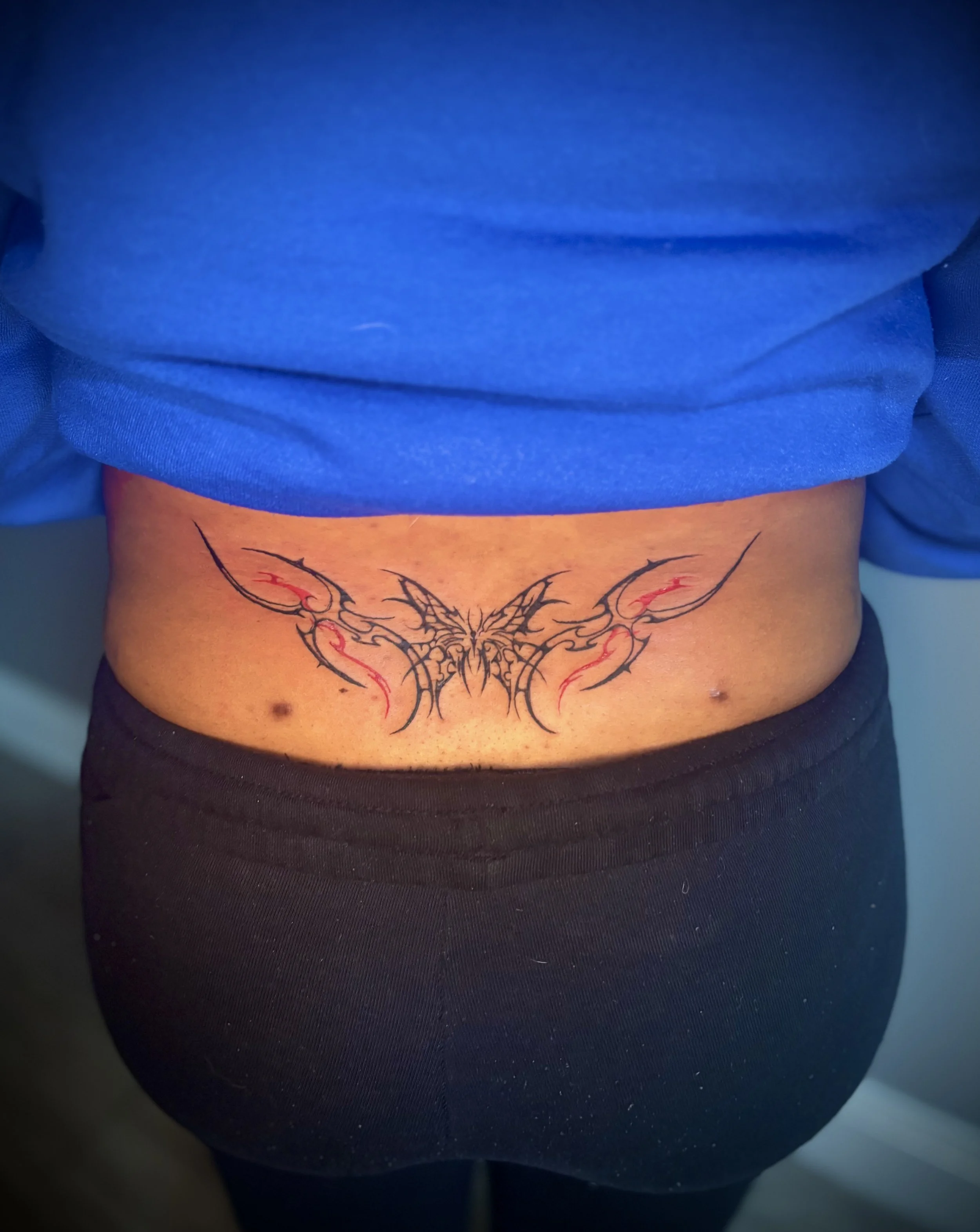Close-up of a person's lower back with a tattoo of a symmetrical, black and red butterfly on the stomach. Cybersigilism style