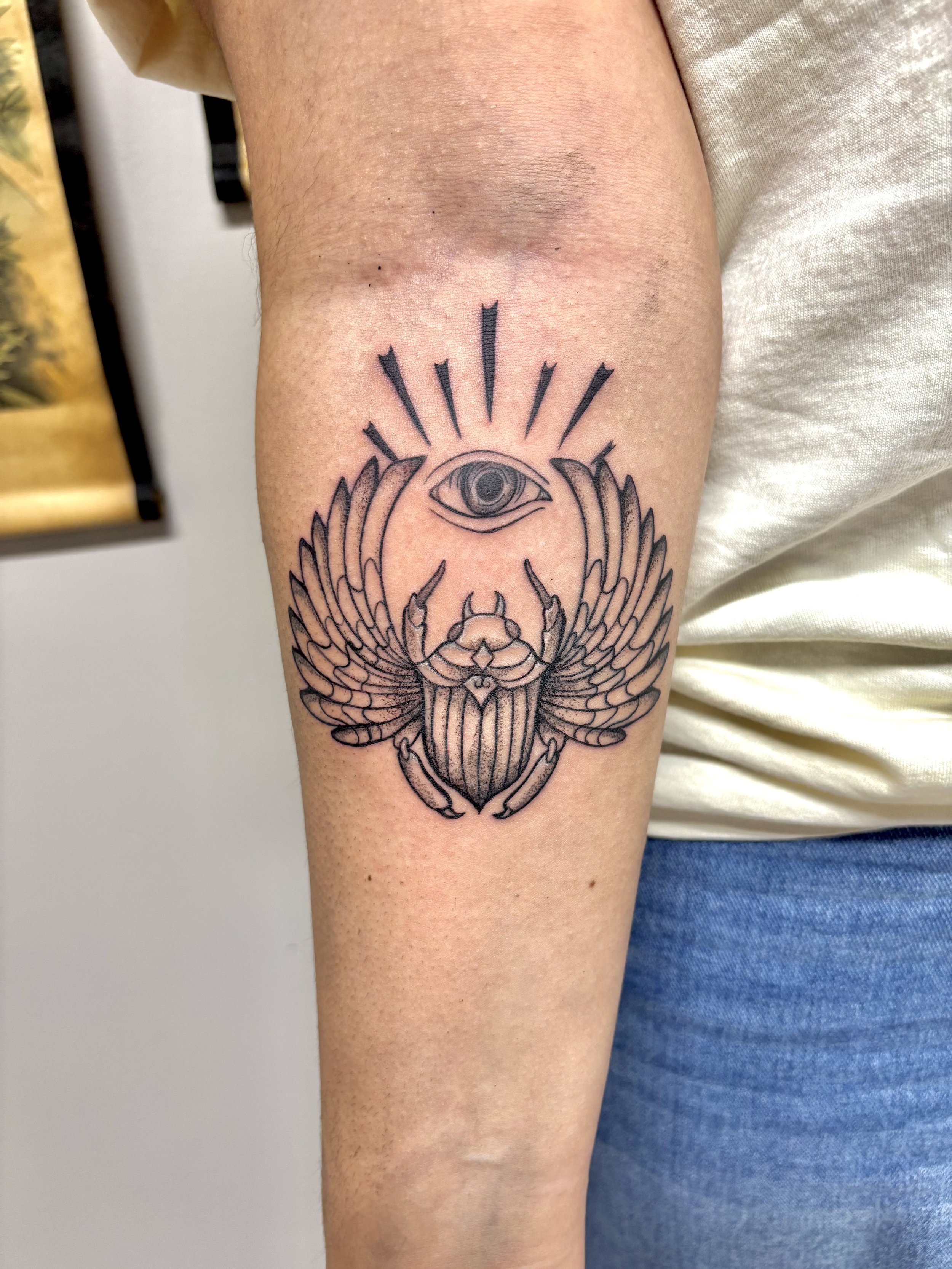 Tattoo of an eye above a scarab beetle with wings on a person's upper arm.