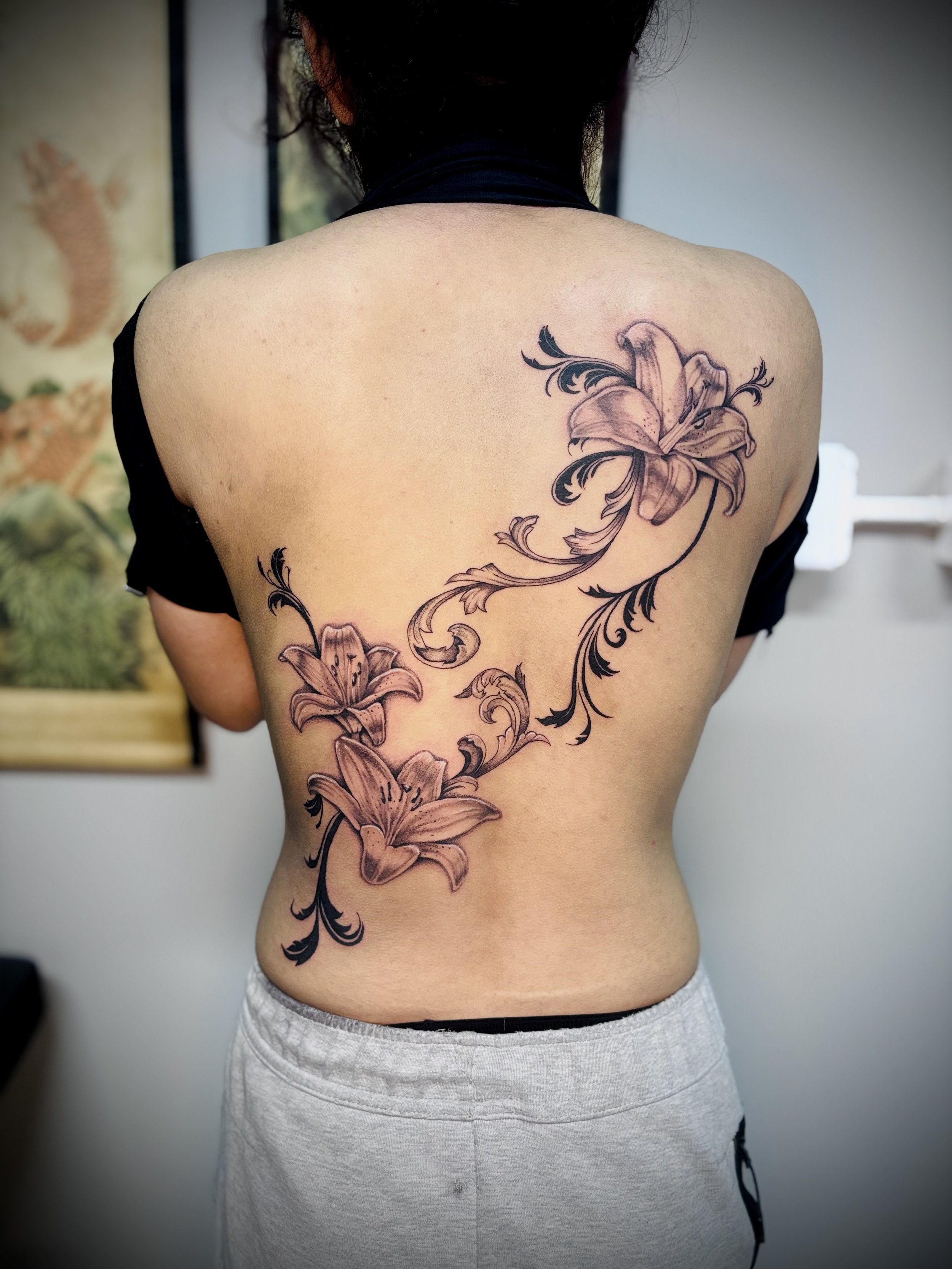 Back view of a person with a large floral tattoo on their back, wearing light gray sweatpants and a black top around their neck. Back tattoo. Realism tattoo. Ornamental tattoo. 