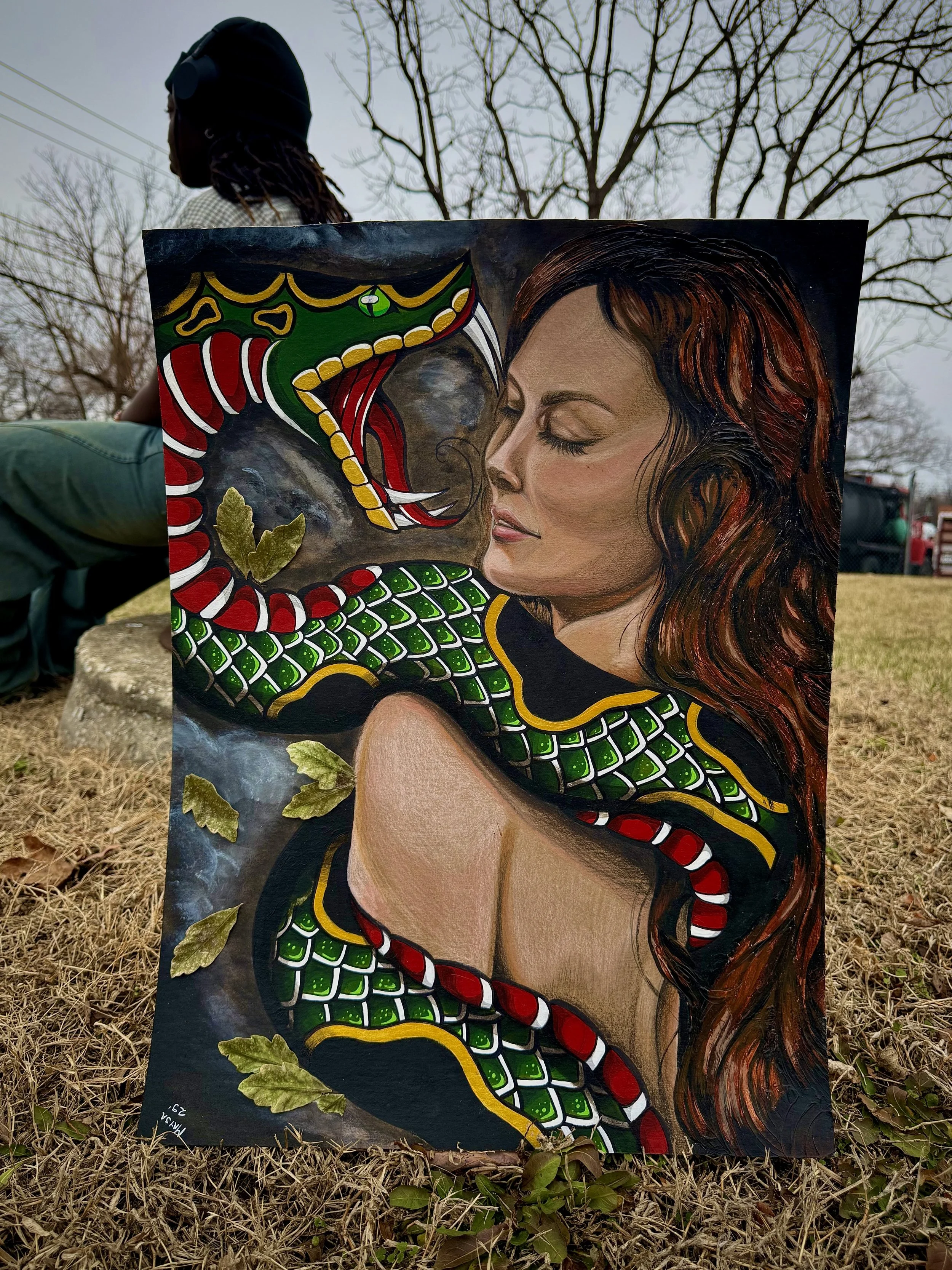 A painting of a woman with closed eyes and long hair, facing a menacing snake with vibrant green, red, and yellow scales and fangs. The painting is held upright outdoors on the ground with dried grass and fallen leaves, and a person with dark hair an