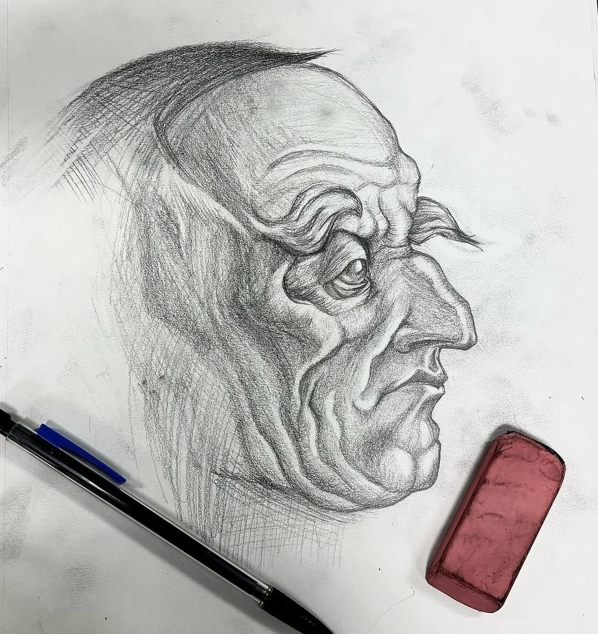 A detailed pencil sketch of an old man's face in profile view, showing wrinkles and expressive features, alongside a black pen and a pink eraser on a white surface.