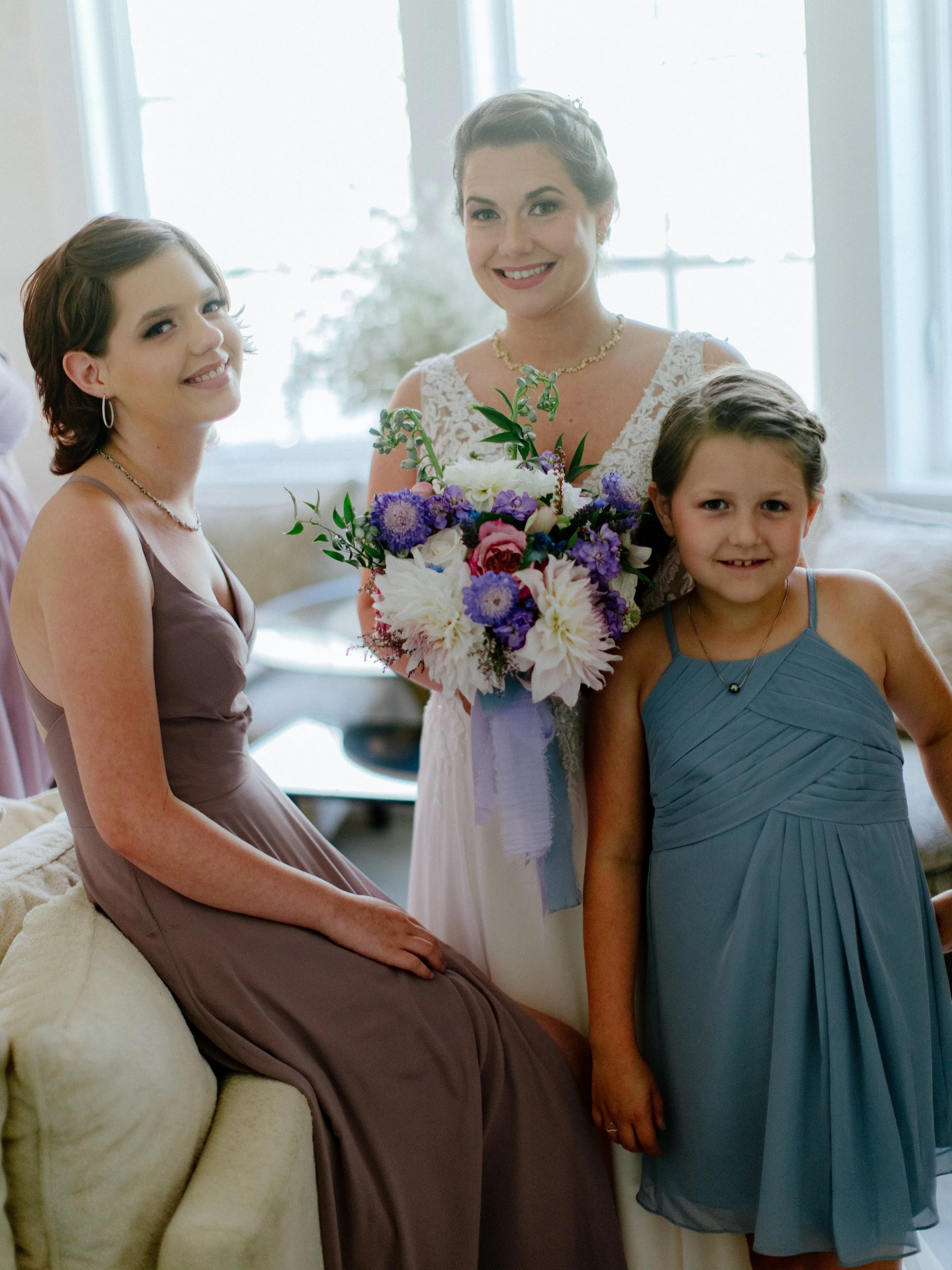 A woman in a wedding dress holding a bouquet of flowers, standing with two younger females, one seated and one standing, all smiling indoors.