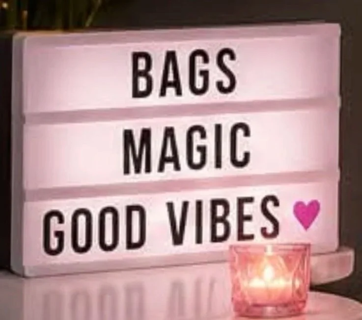 A pink lightbox sign with black letters reading "BAGS MAGIC GOOD VIBES," and a pink heart symbol below. A lit candle in a pink holder is placed in front of the sign.