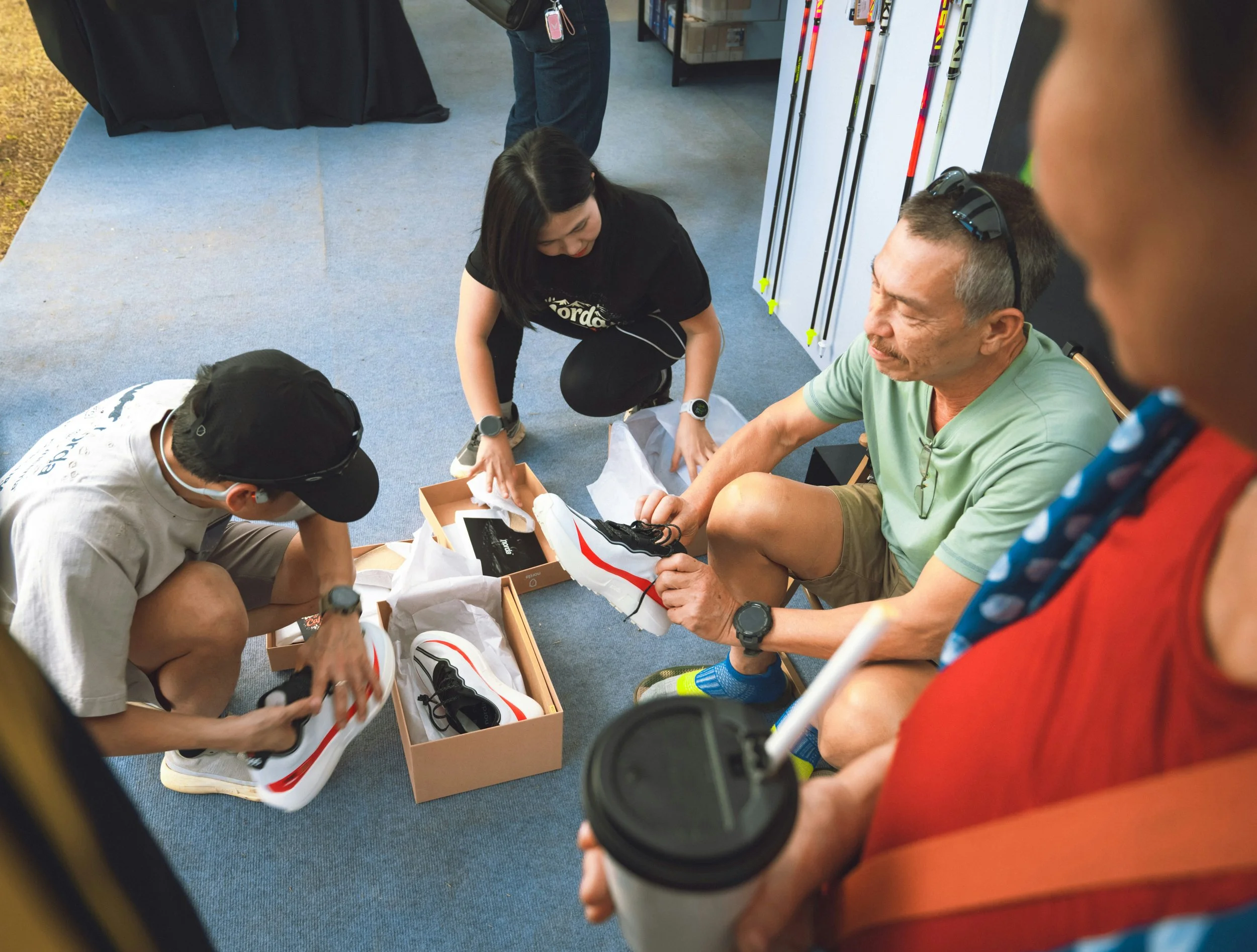 People sitting on the floor in a circle, exchanging shoes, inside a store with sports equipment displayed on the wall.