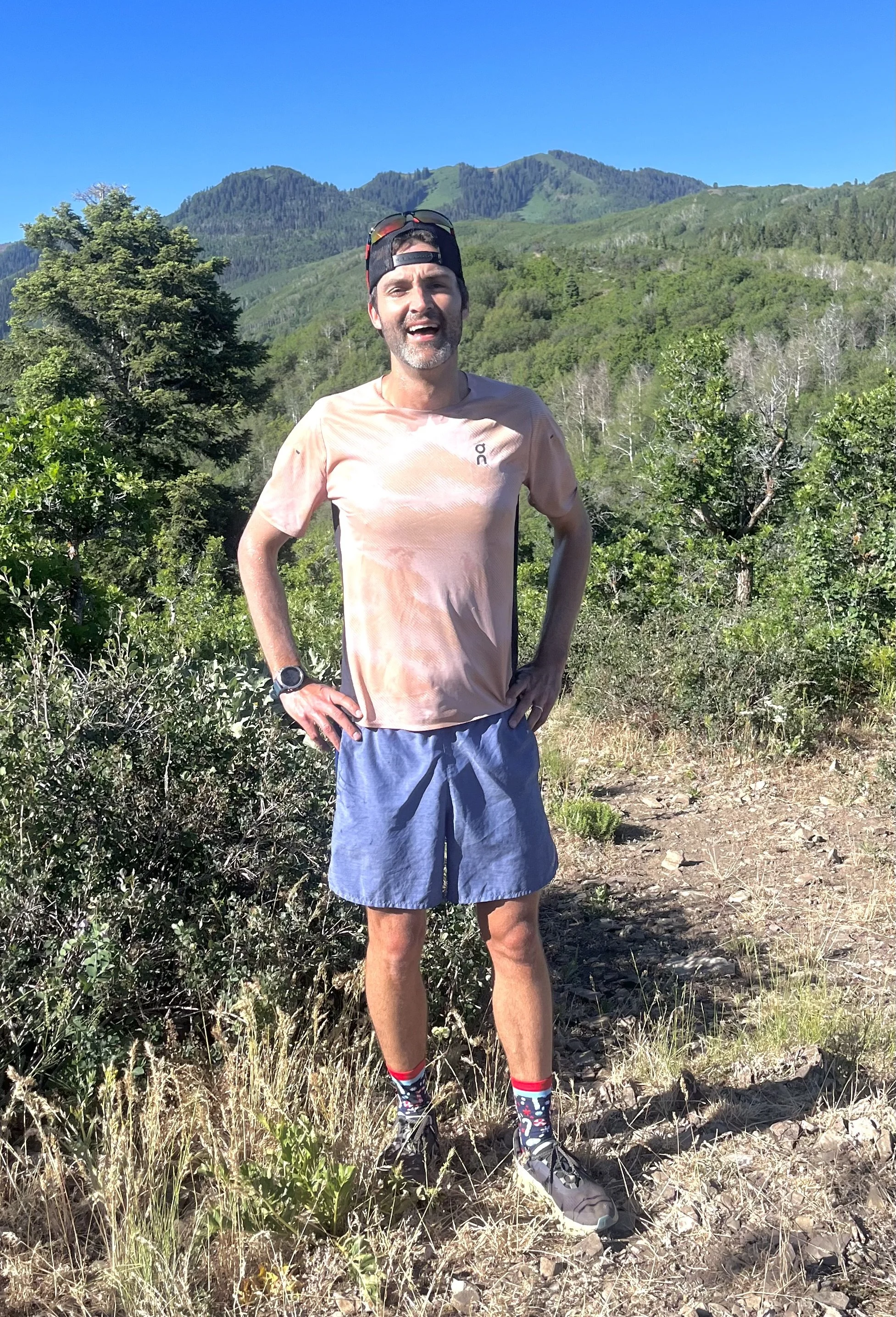 A man standing outdoors on a trail in a mountainous area with green trees and a clear blue sky, wearing a peach-colored t-shirt, blue shorts, running shoes, and sunglasses on his head.