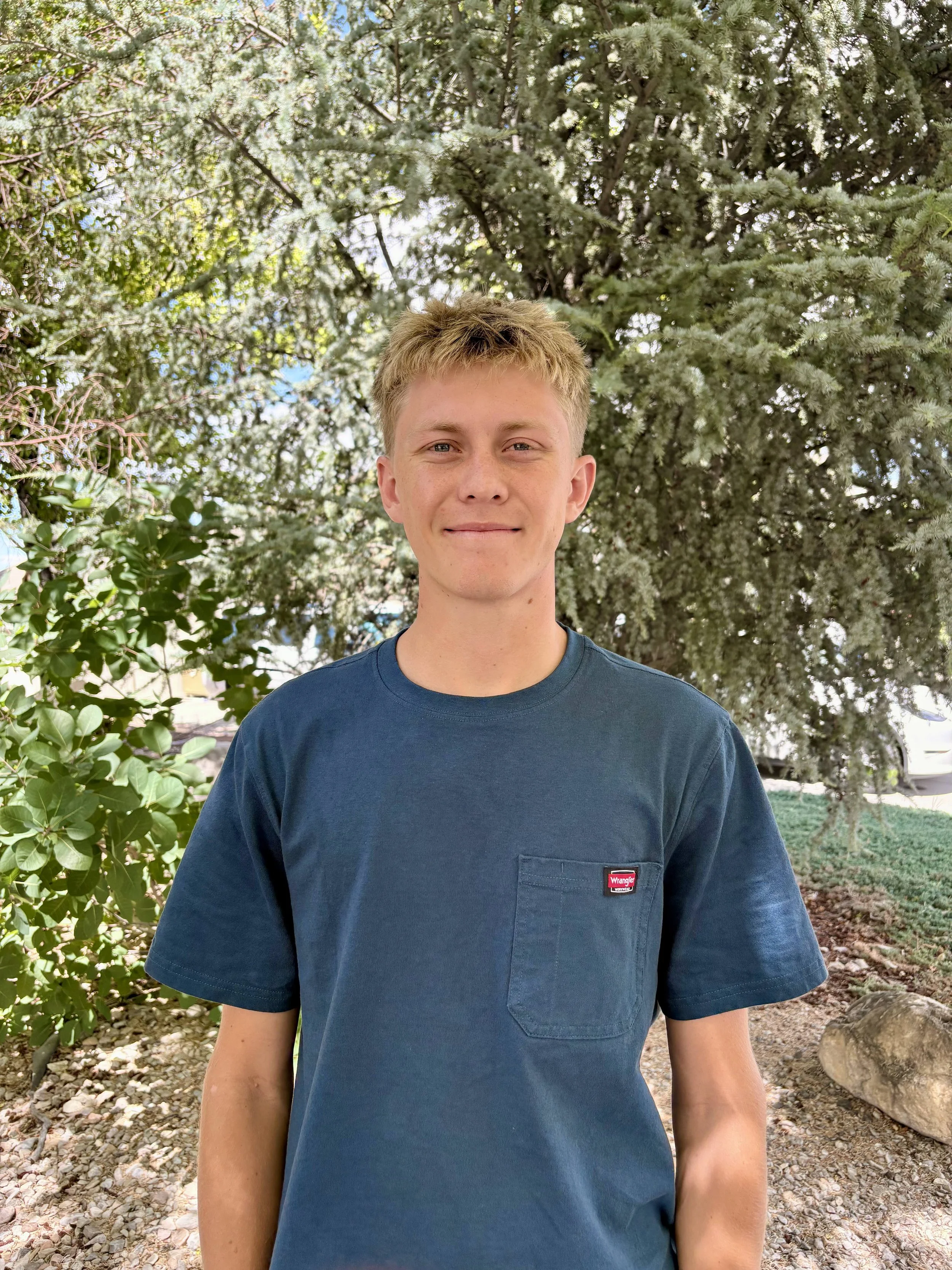 A young man with short blond hair wearing a dark blue T-shirt standing outdoors in front of a background of green trees and plants.