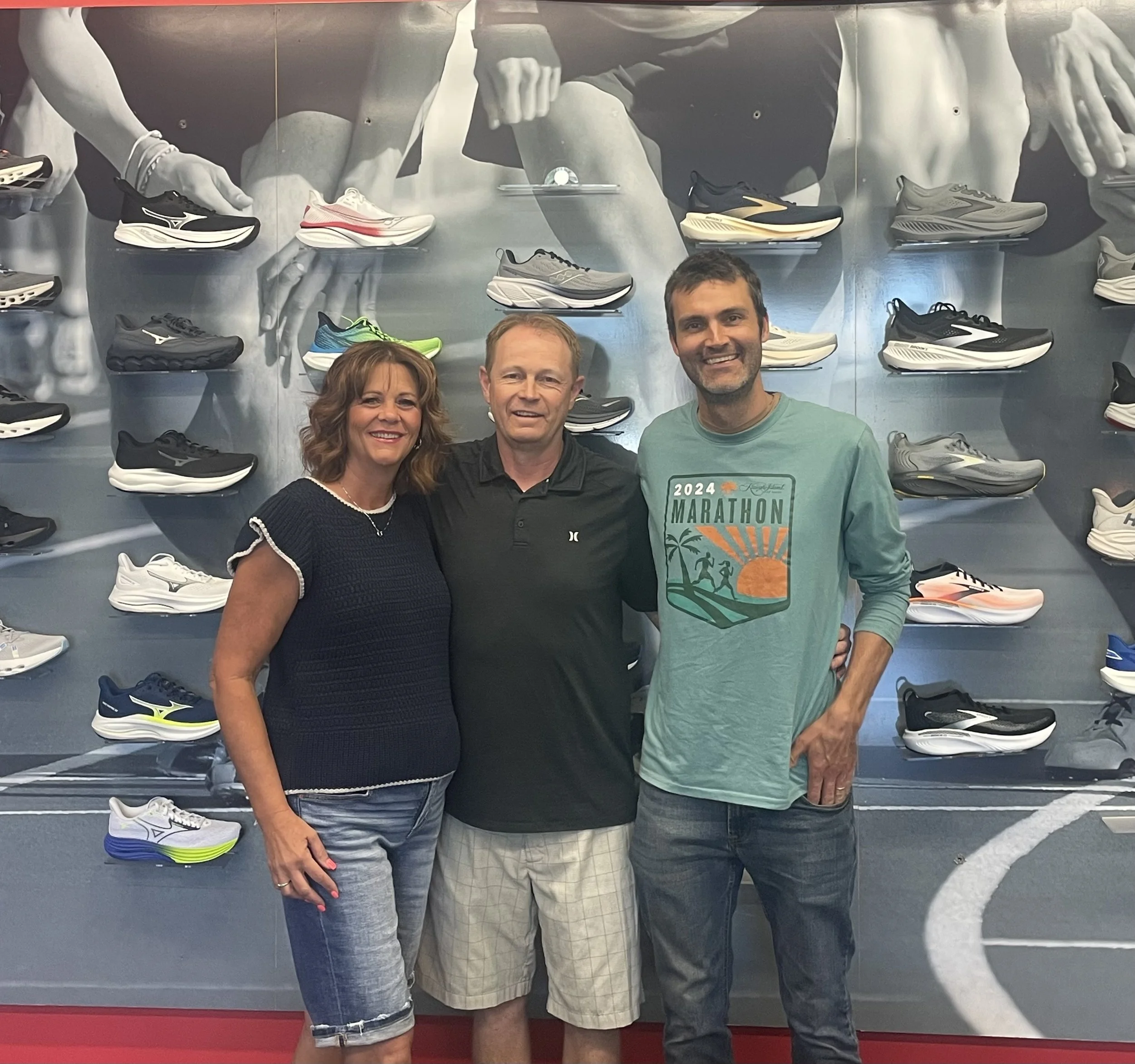 Three people standing side by side, smiling, in front of a display wall of athletic shoes.