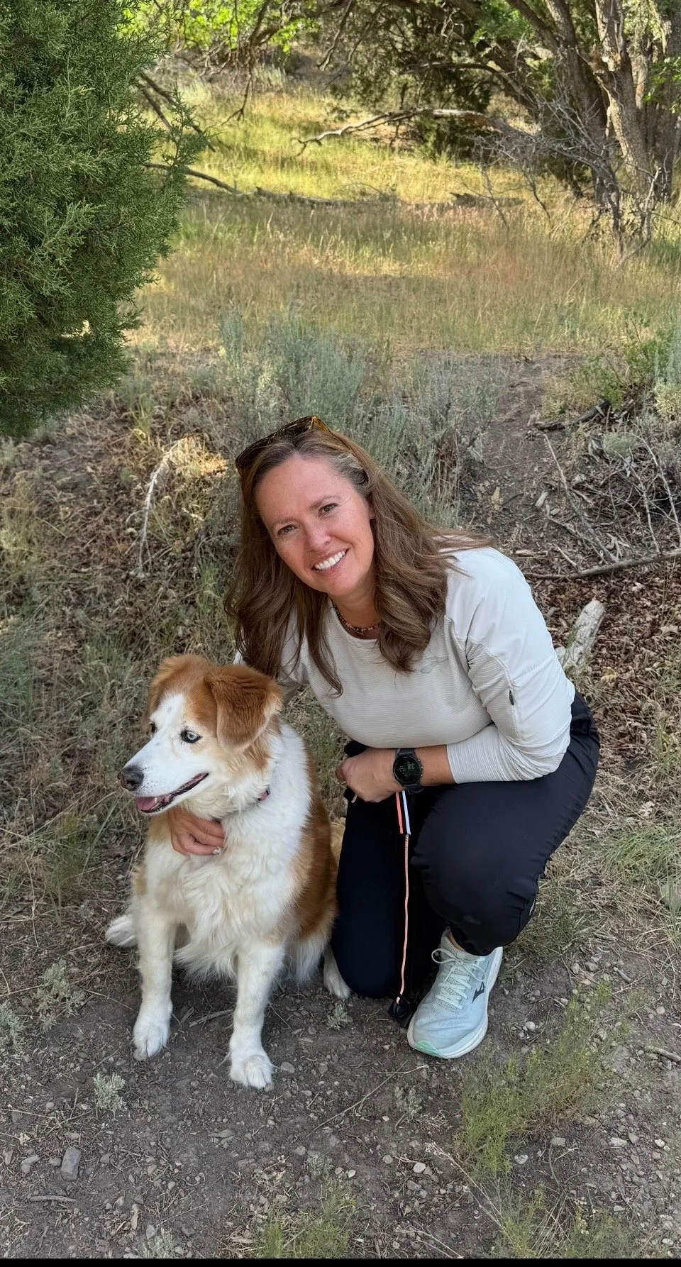 A woman smiling and squatting on the ground next to a border collie dog in a natural outdoor setting with trees and grass.