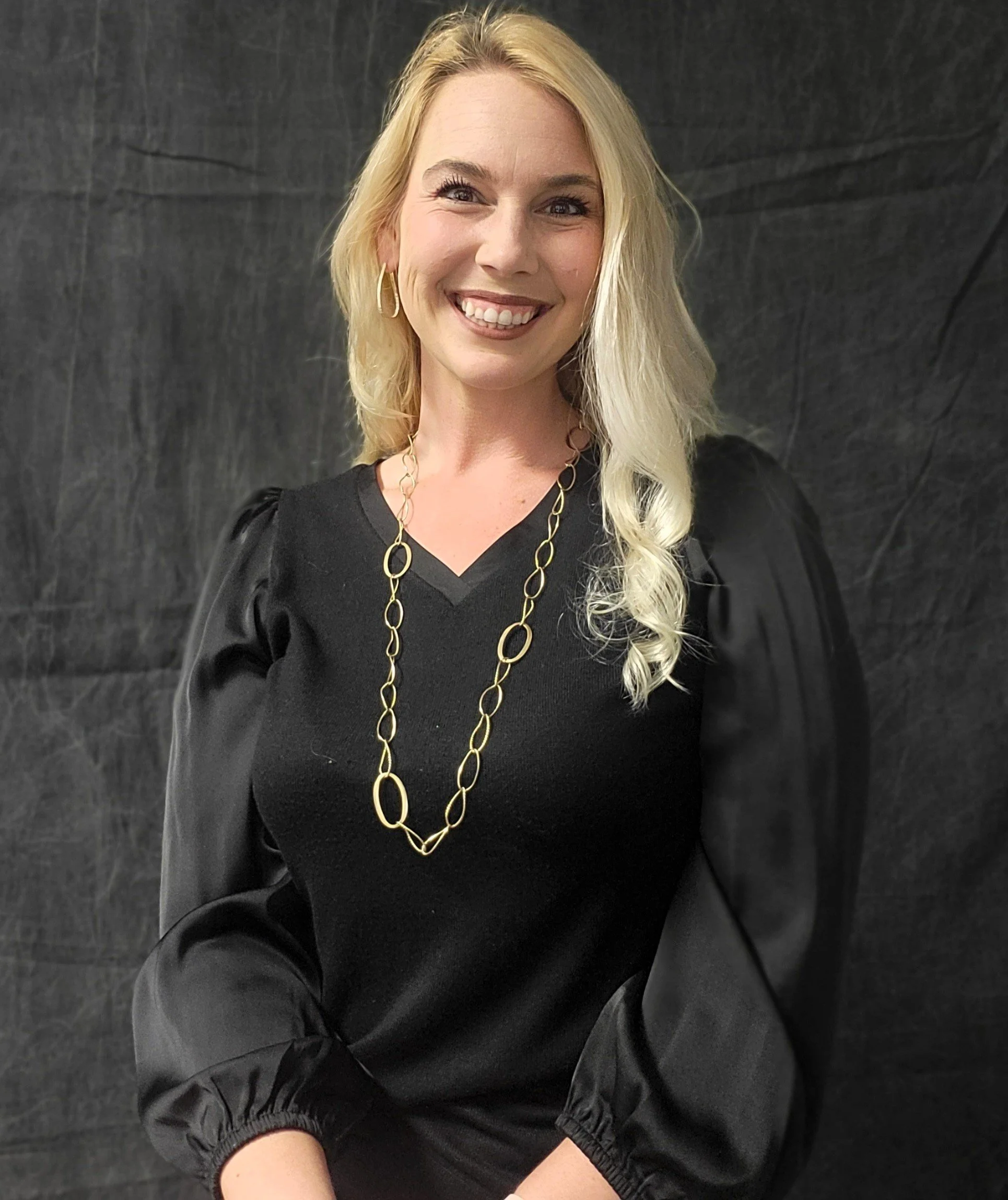 A woman with long blonde hair smiling, wearing a black top, large gold hoop earrings, and a long gold chain necklace, against a dark textured background.