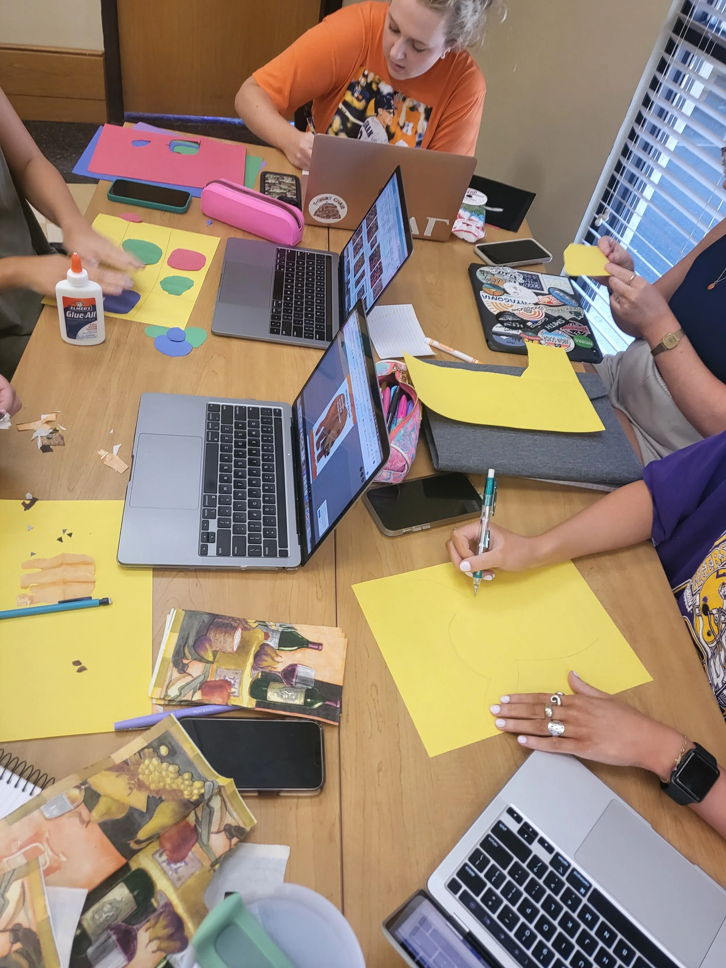 People working on arts and crafts projects at a table with laptops, paper cutouts, glue, and magazines.