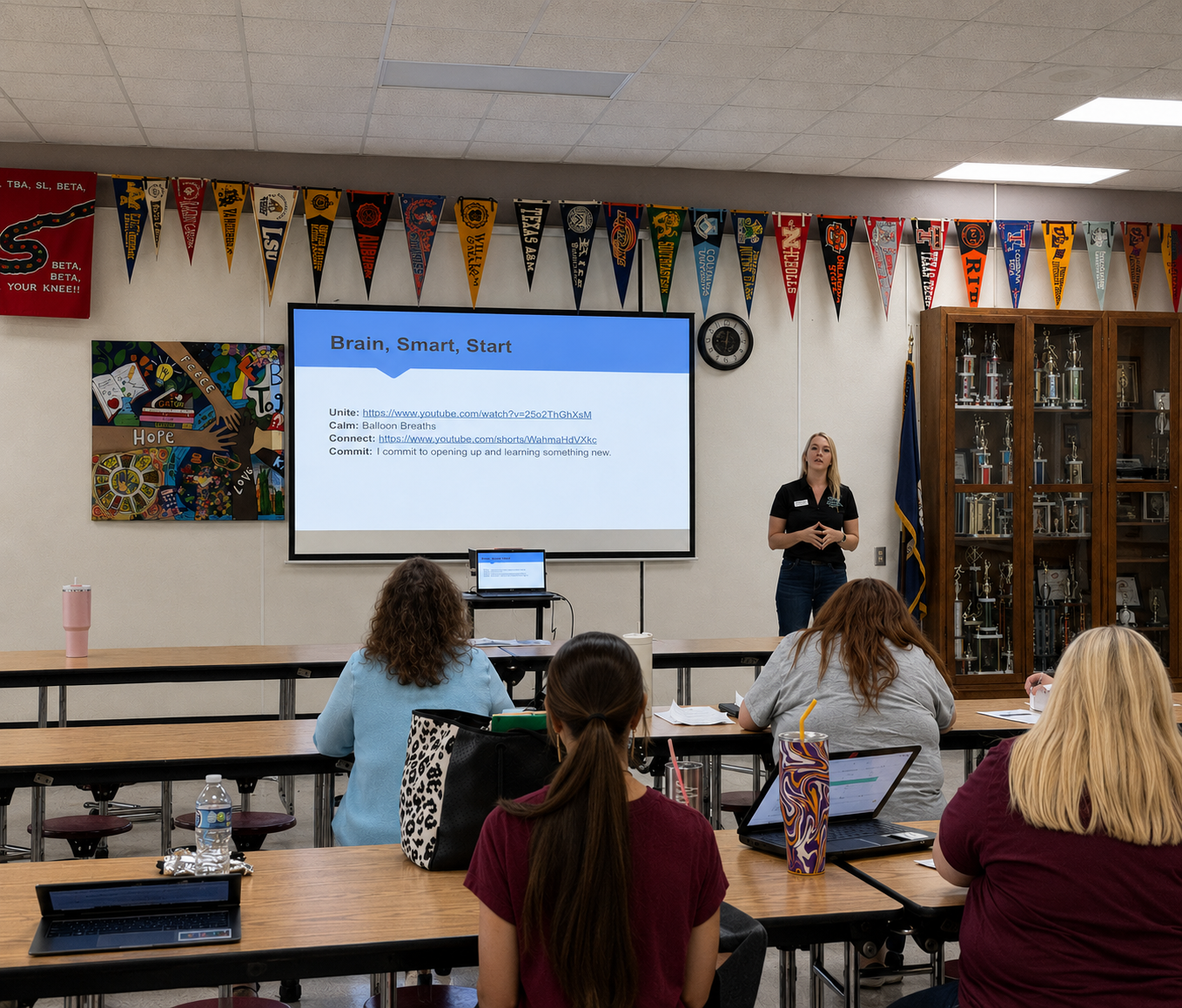 A classroom with five women seated at wooden tables, facing a presenter standing in front of a large screen displaying a presentation titled "Brain, Smart, Start." The classroom is decorated with colorful banners and a trophy cabinet.