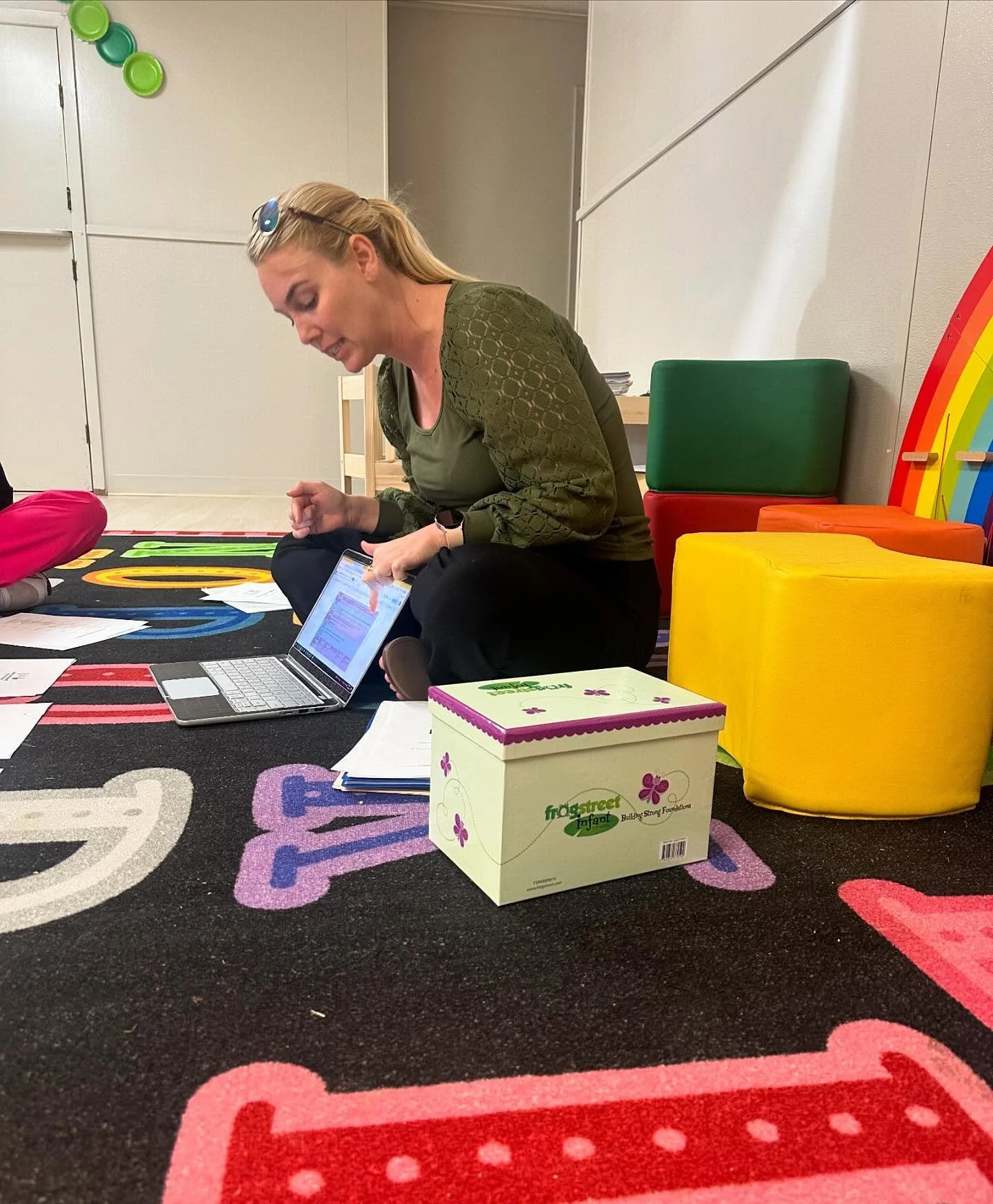 A woman sitting on a colorful children's rug in a room with brightly colored furniture and papers around her. She is using a tablet, with a box labeled 'Frost Street Infant' nearby.