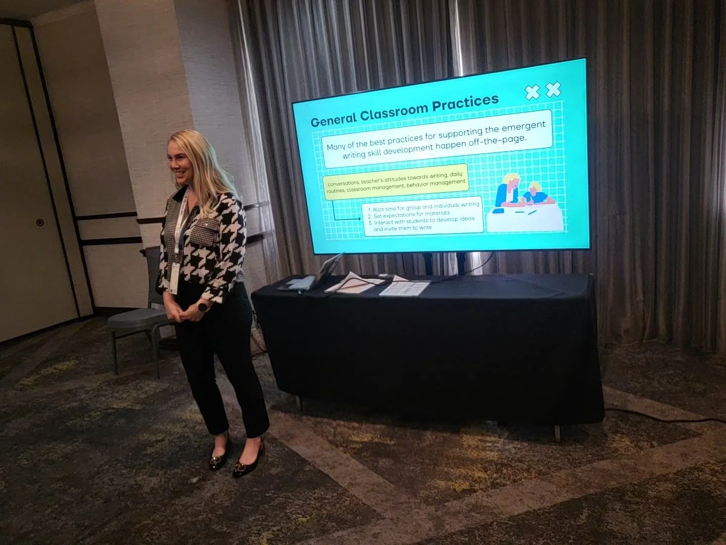 A woman standing next to a table with a large digital screen displaying a presentation titled 'General Classroom Practices.' The woman is smiling, wearing a black and white patterned blazer, black pants, and black heels. The presentation on the screen includes text about supporting emergent writing skills
