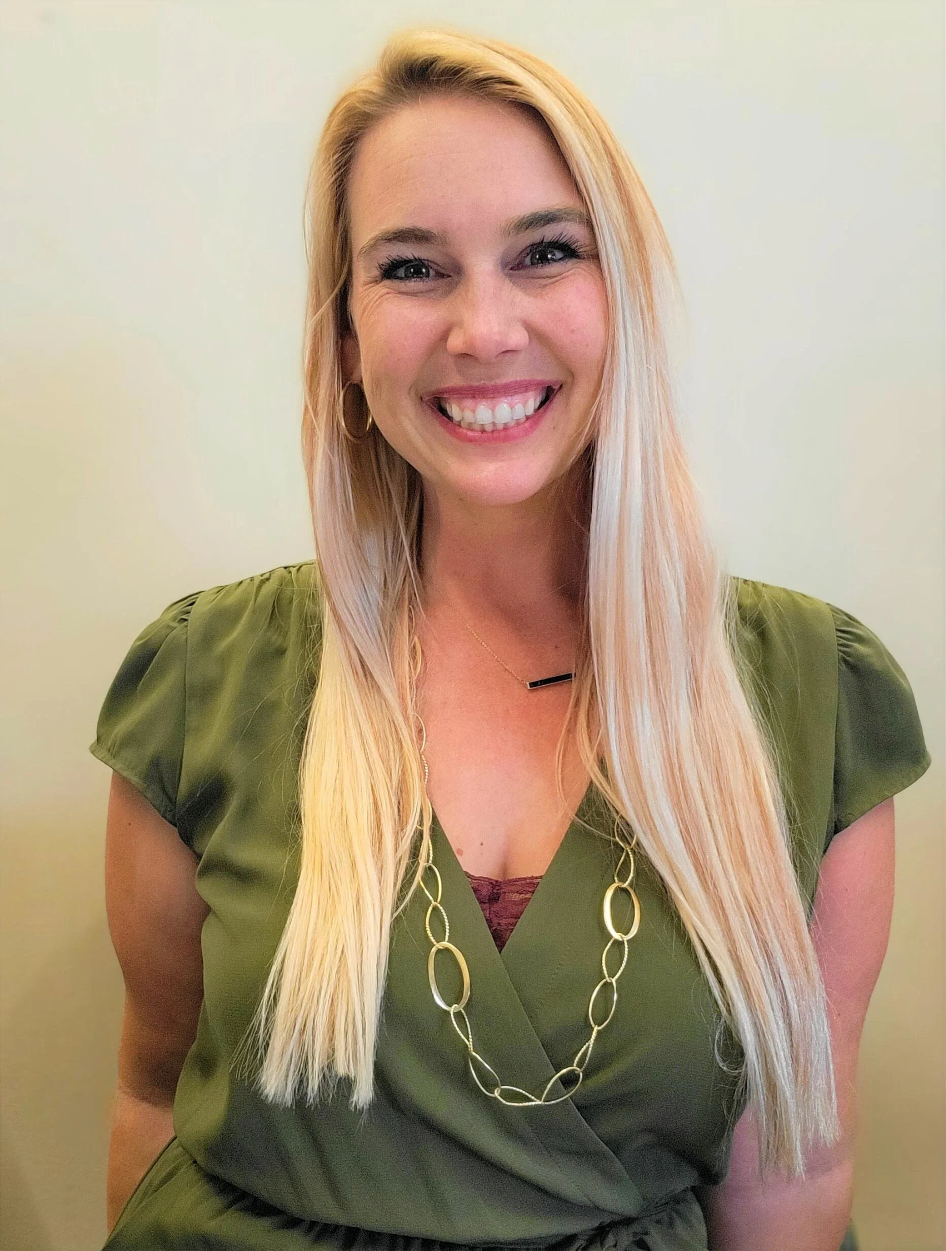 A woman with long blonde hair smiling, wearing a green blouse with puffed sleeves, hoop earrings, and a long chain necklace, standing against a plain light-colored wall.