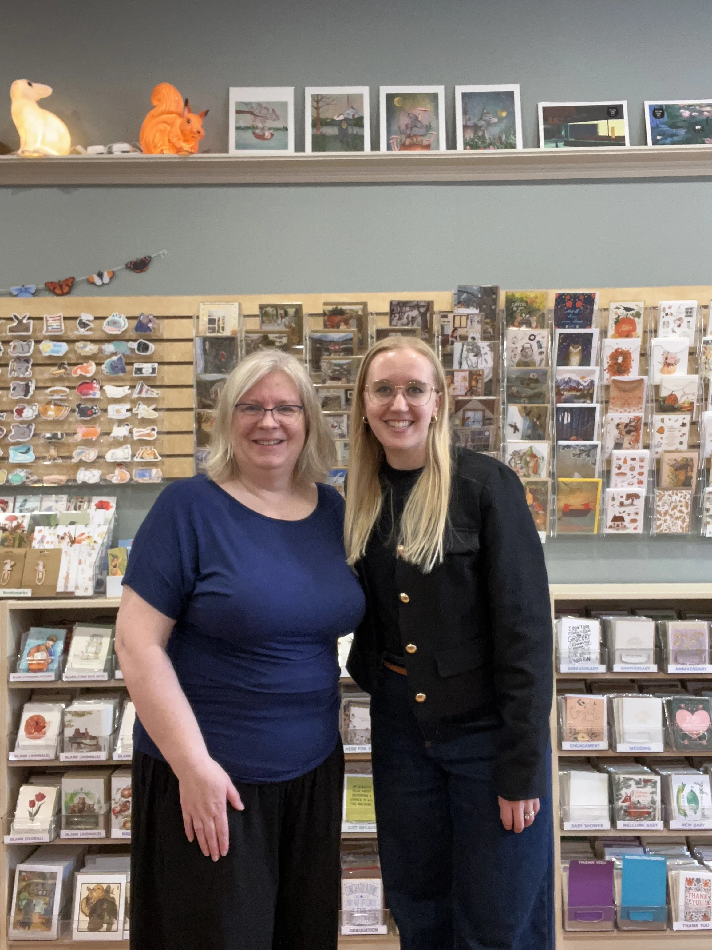 Two women standing in front of shelves filled with greeting cards, smiling at the camera. The woman on the left has blonde hair and glasses, wearing a blue top. The woman on the right has long blonde hair, glasses, and is wearing a dark jacket with gold buttons.