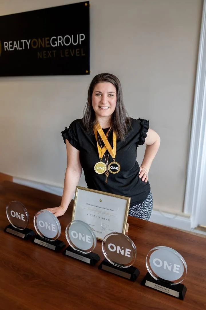 A woman with dark hair smiling, wearing black top with ruffled sleeves and patterned pants, standing behind a wooden table displaying multiple awards and a framed certificate. She is wearing medals around her neck. In the background, a sign on the wa