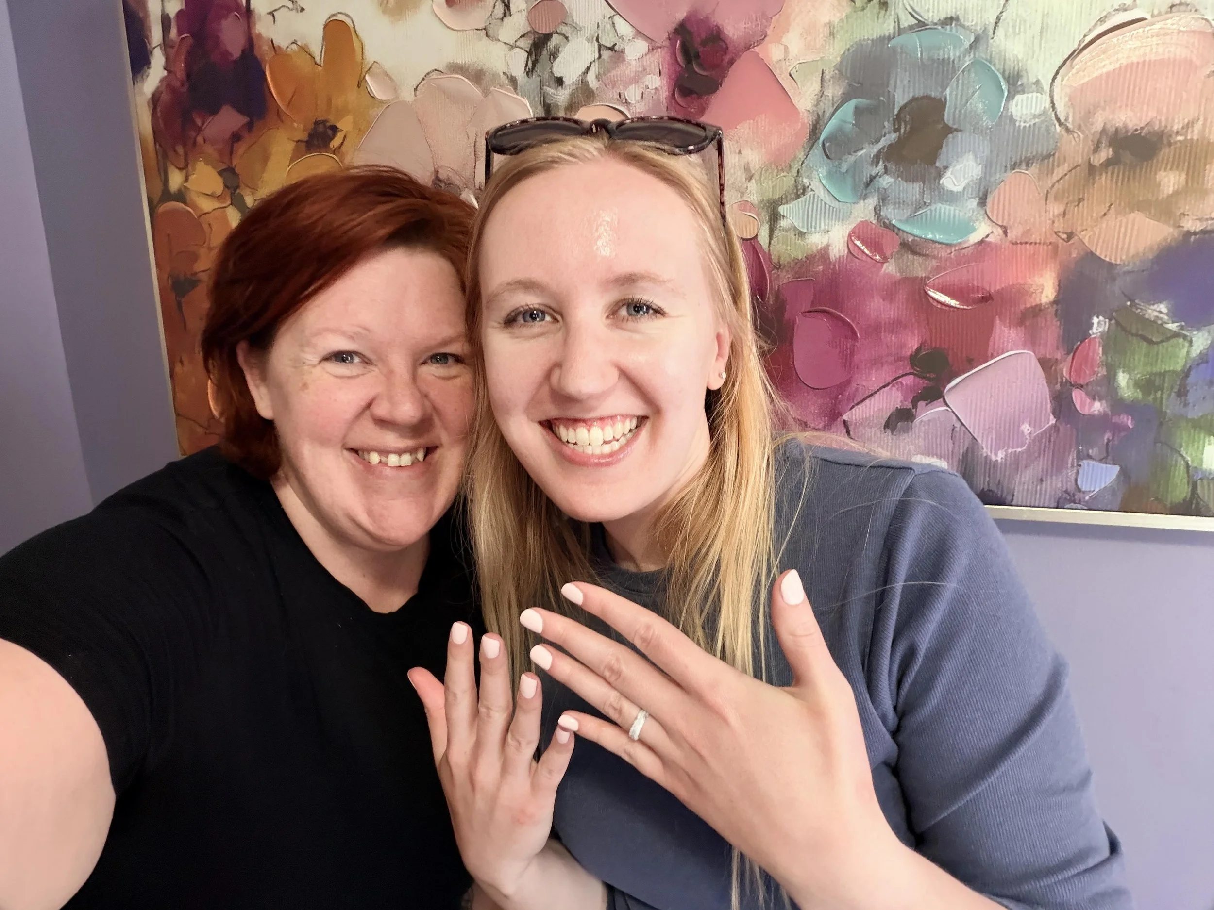 Close-up of two women smiling, showing their rings, with a colorful abstract painting in the background.