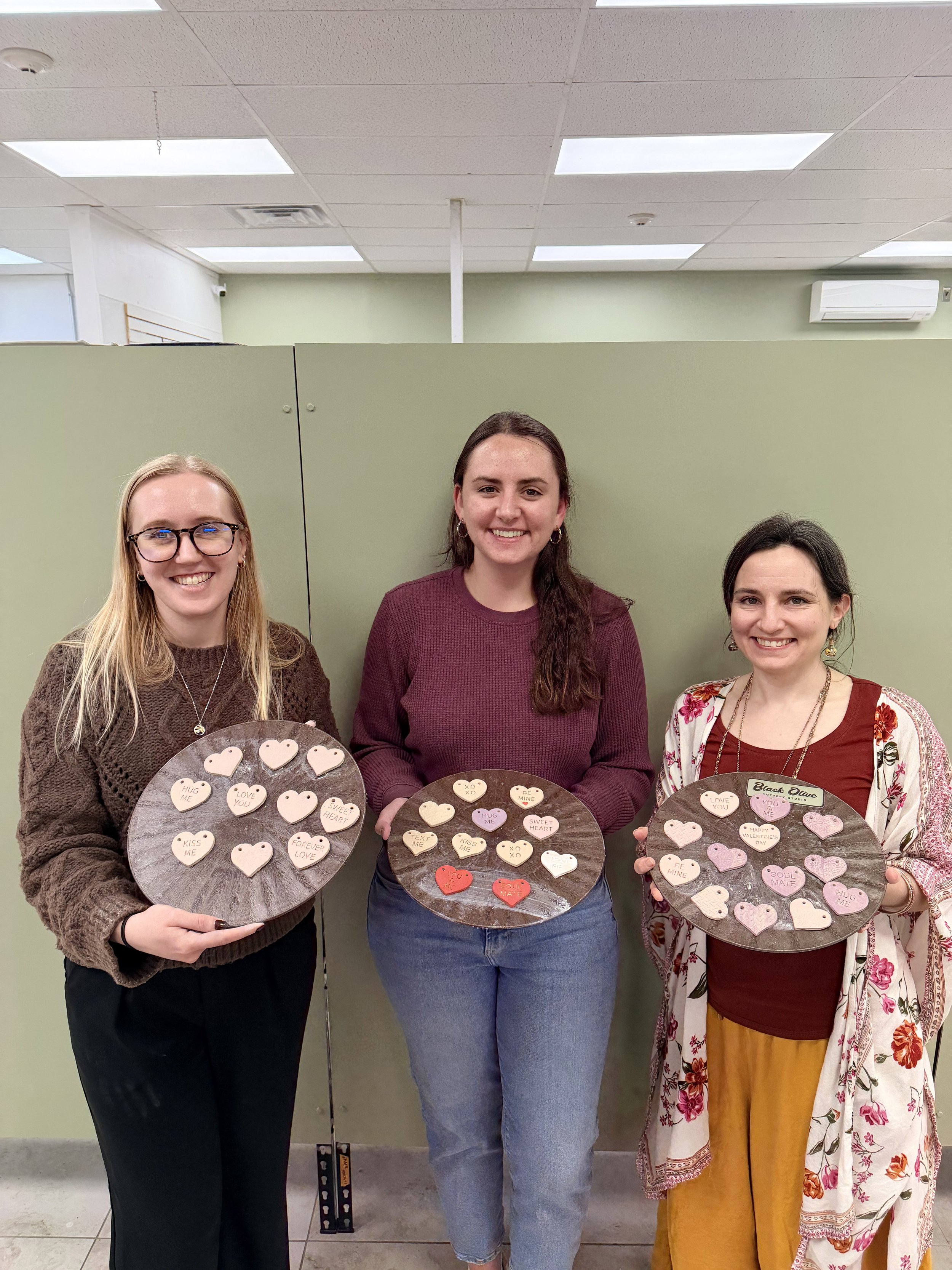 Three women standing indoors behind a greenish partition, each holding a tray with decorated cookies in heart shapes with messages, smiling at the camera.