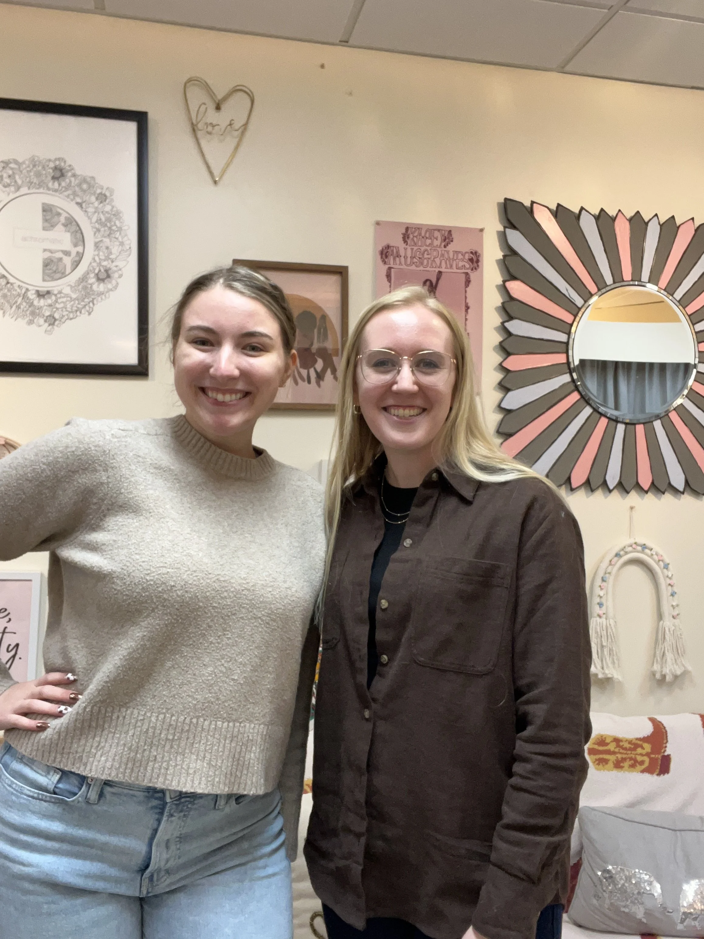 Two women standing close together, smiling at the camera, in an indoor setting with wall art and decorations behind them, including a heart-shaped wire art and mirrors.