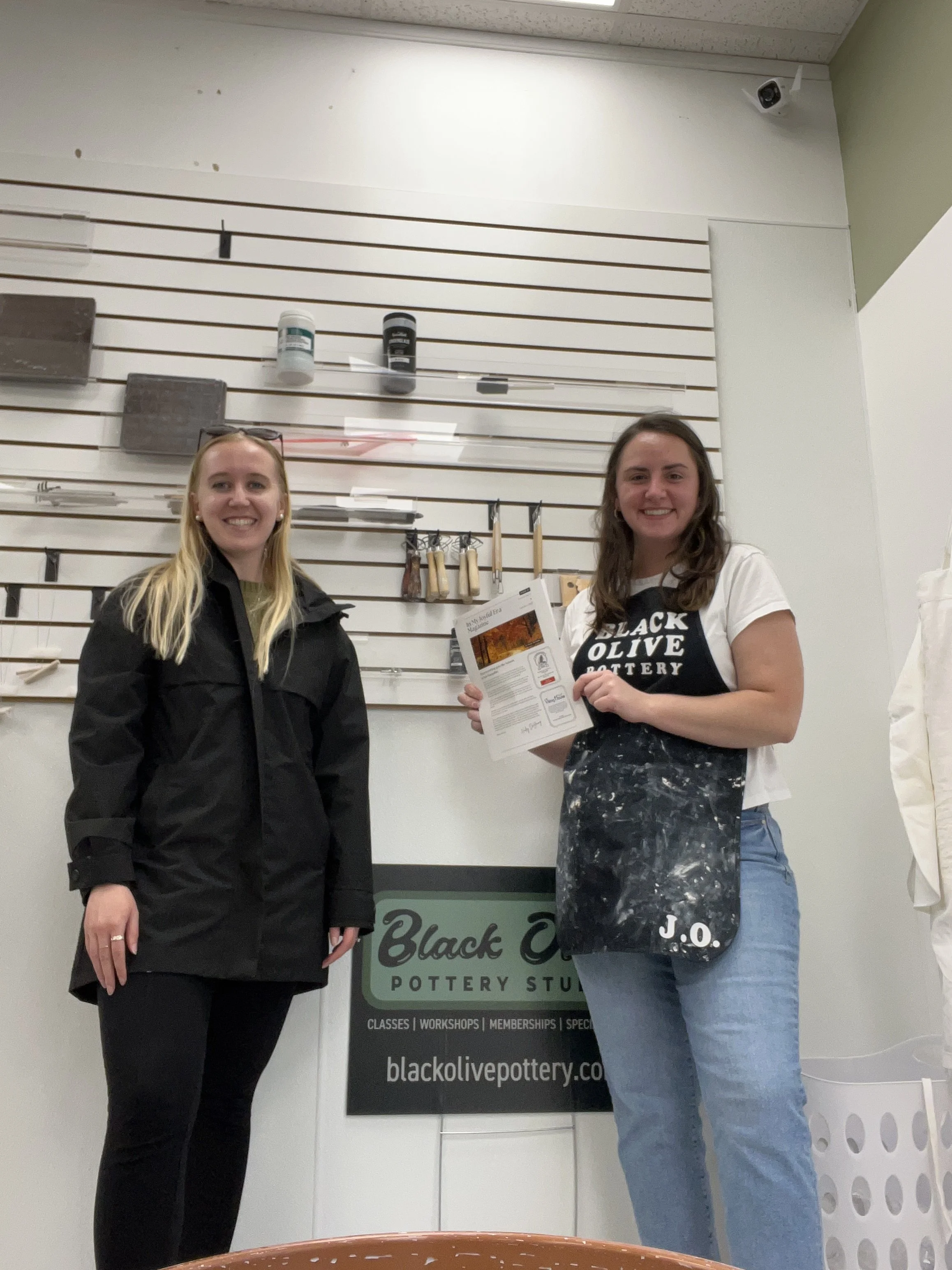 Two women standing in front of a wall display with pottery tools and a sign that reads 'Black Olive Pottery Studio'. One woman is holding a brochure and both are smiling.