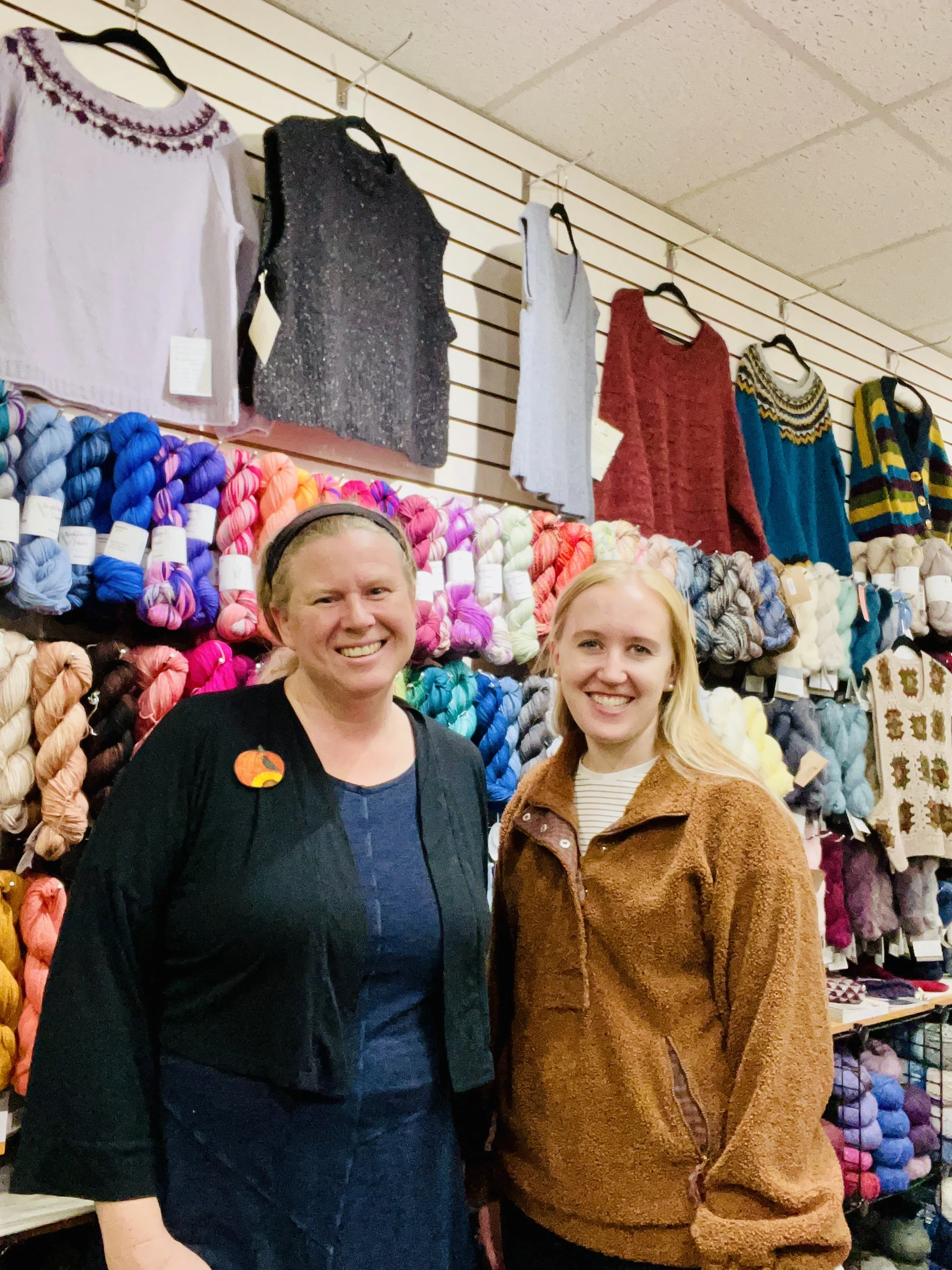 Two smiling women standing in front of a wall display of colorful yarn skeins and sweaters.