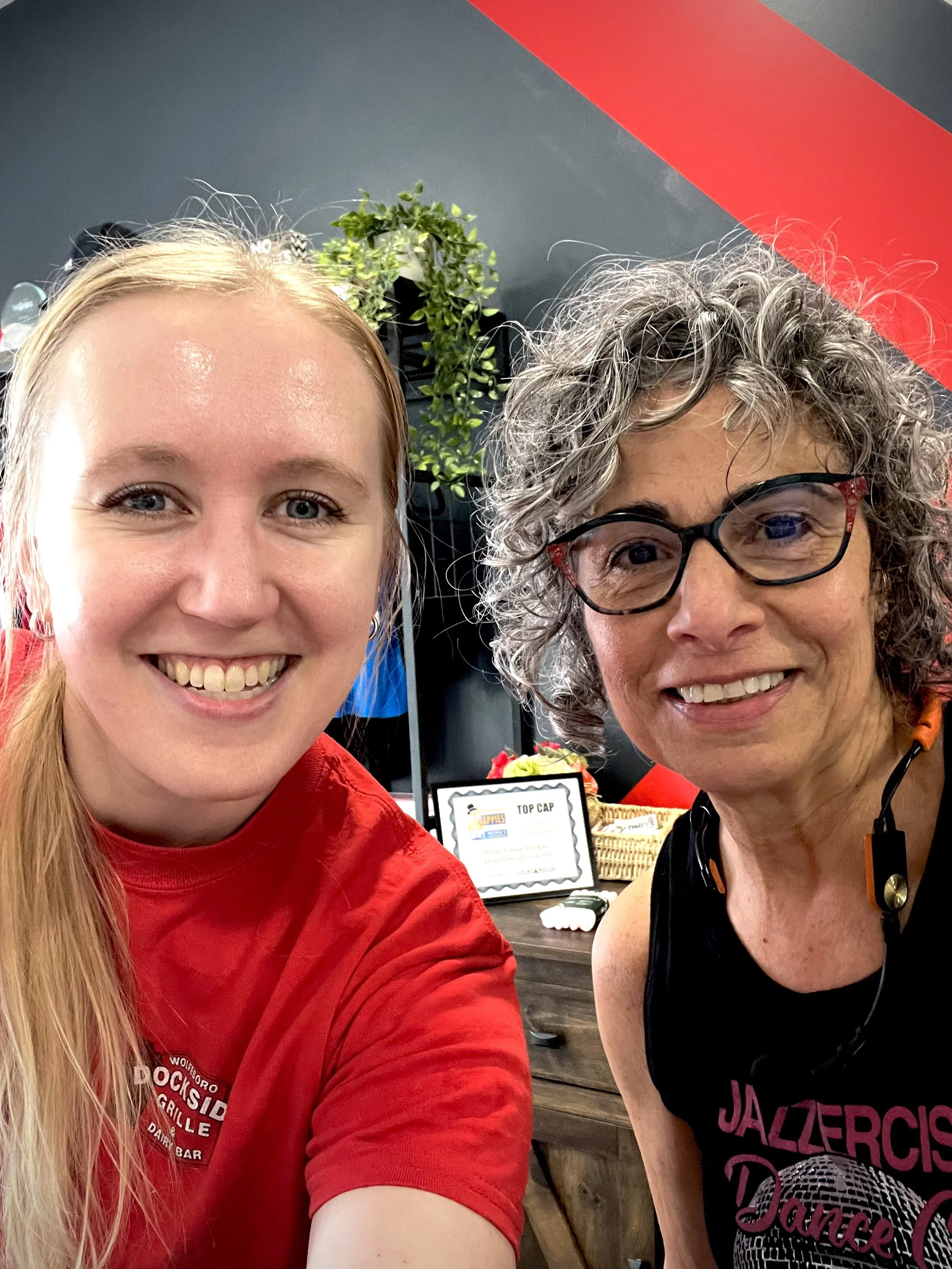 Two women smiling, taking a selfie in a restaurant or cafe with a grey and red wall and a plant in the background.