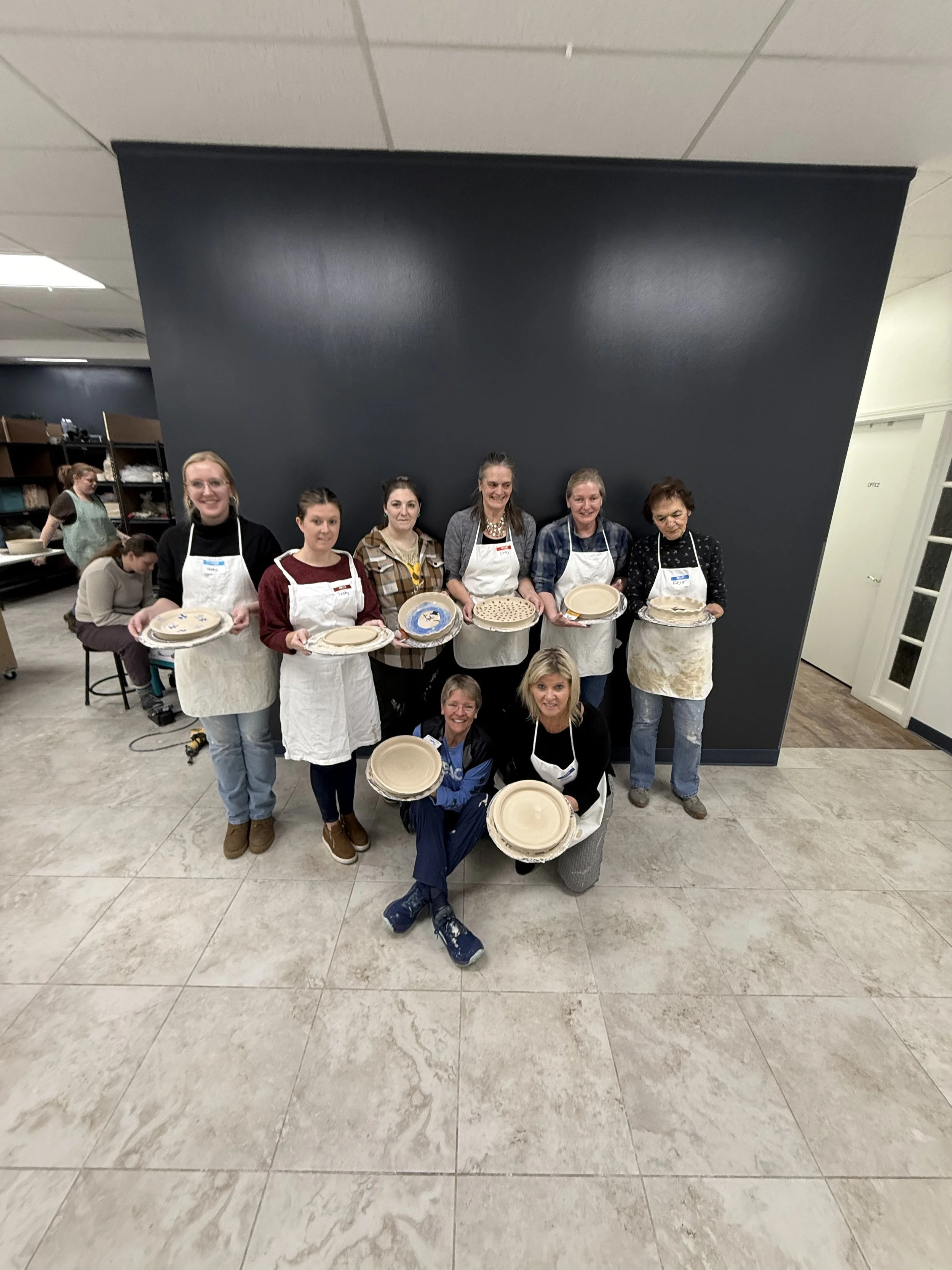 Group of women in apron holding pottery plates in a pottery workshop with others working in background.