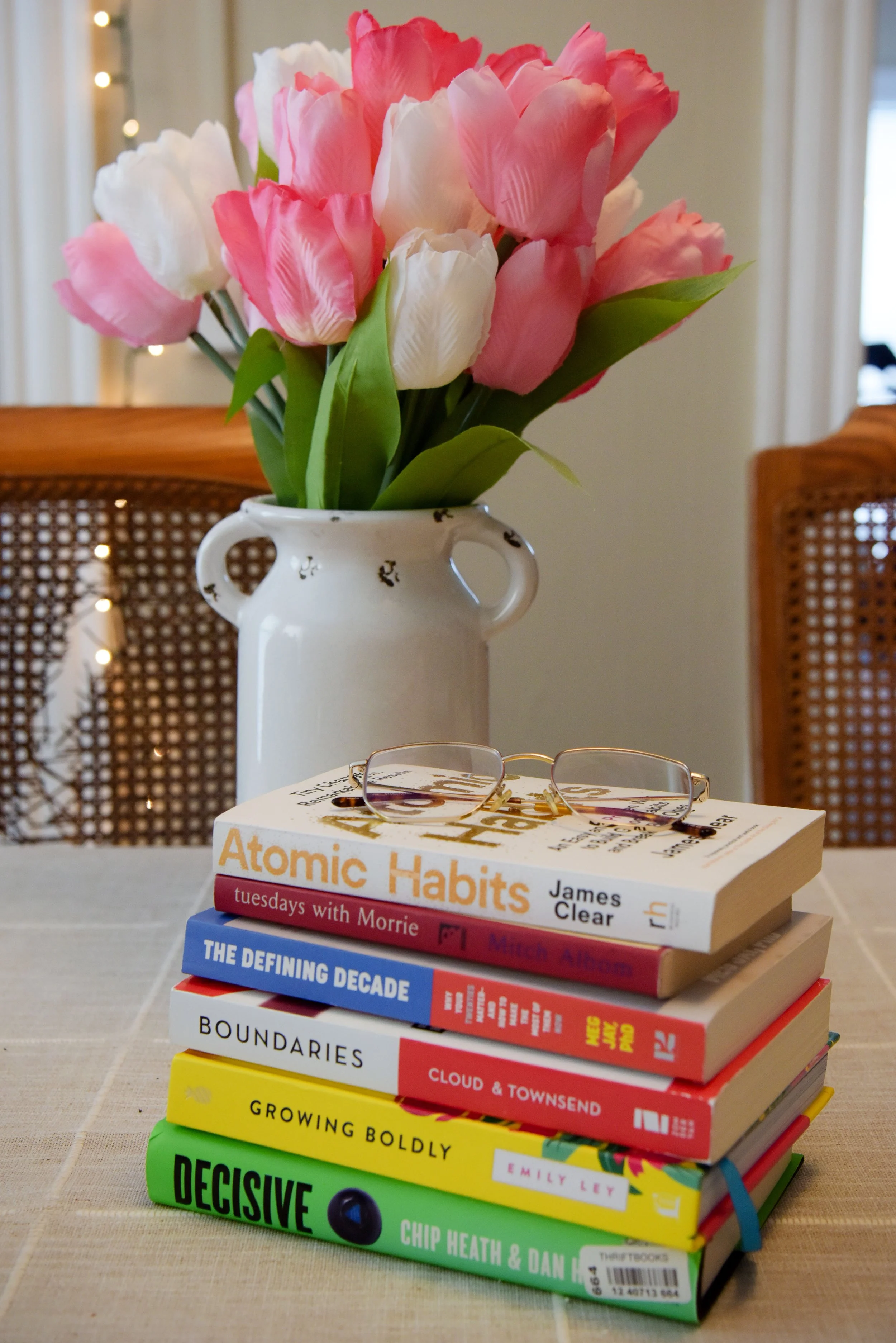 Stack of colorful books titled 'Atomic Habits,' 'Tuesdays with Morrie,' 'The Defining Decade,' 'Boundaries,' 'Cloud & Townsend,' 'Growing Boldly,' 'Decisive' on a table. Behind it is a white vase with pink and white flowers, and a pair of glasses on top of the books.